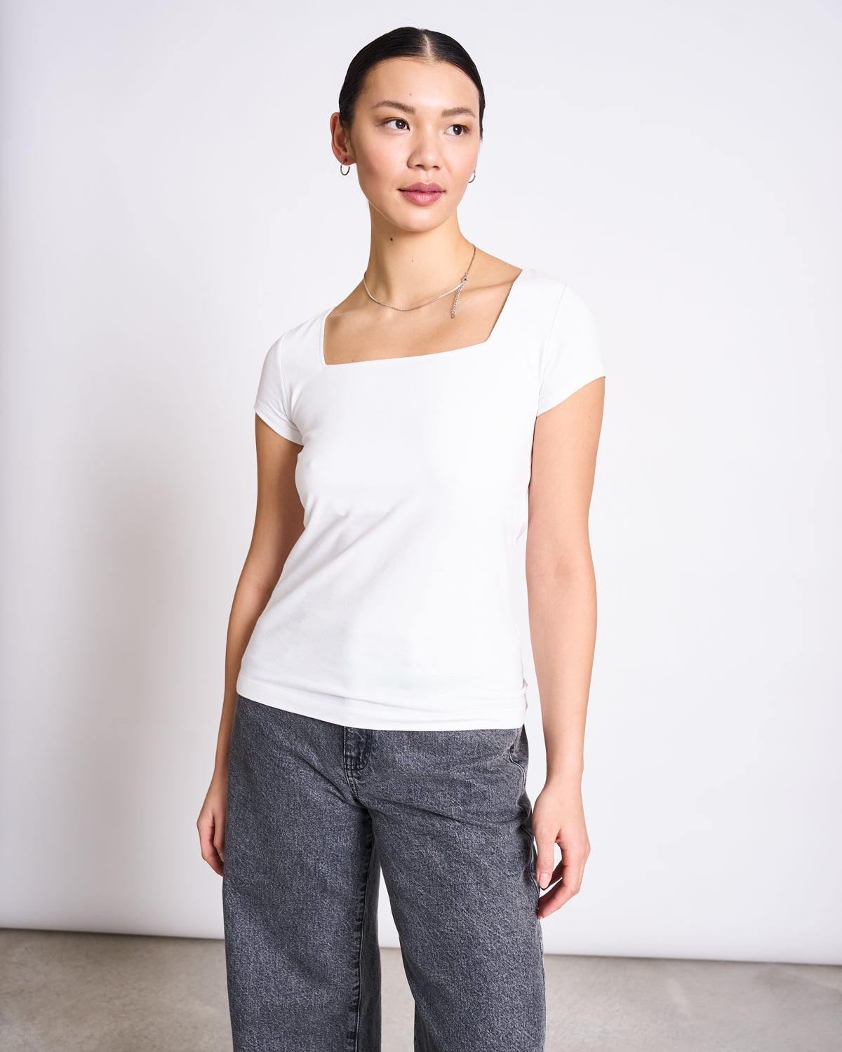 A woman stands against a plain white background, wearing the jan-n-june T-SHIRT ARIMA WHITE GOTS—a fitted square neck short-sleeve top in super soft fabric—paired with loose gray jeans. Her hair is tied back and she gazes gently to the side.