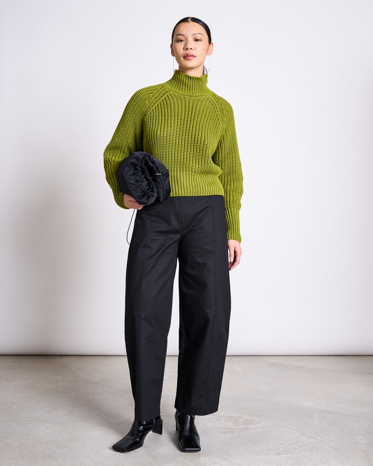 A person stands against a white wall, wearing the jan-n-june JUMPER OLA MOSSY PEAR GOTS, loose black trousers, and black ankle boots, holding a black clutch bag.