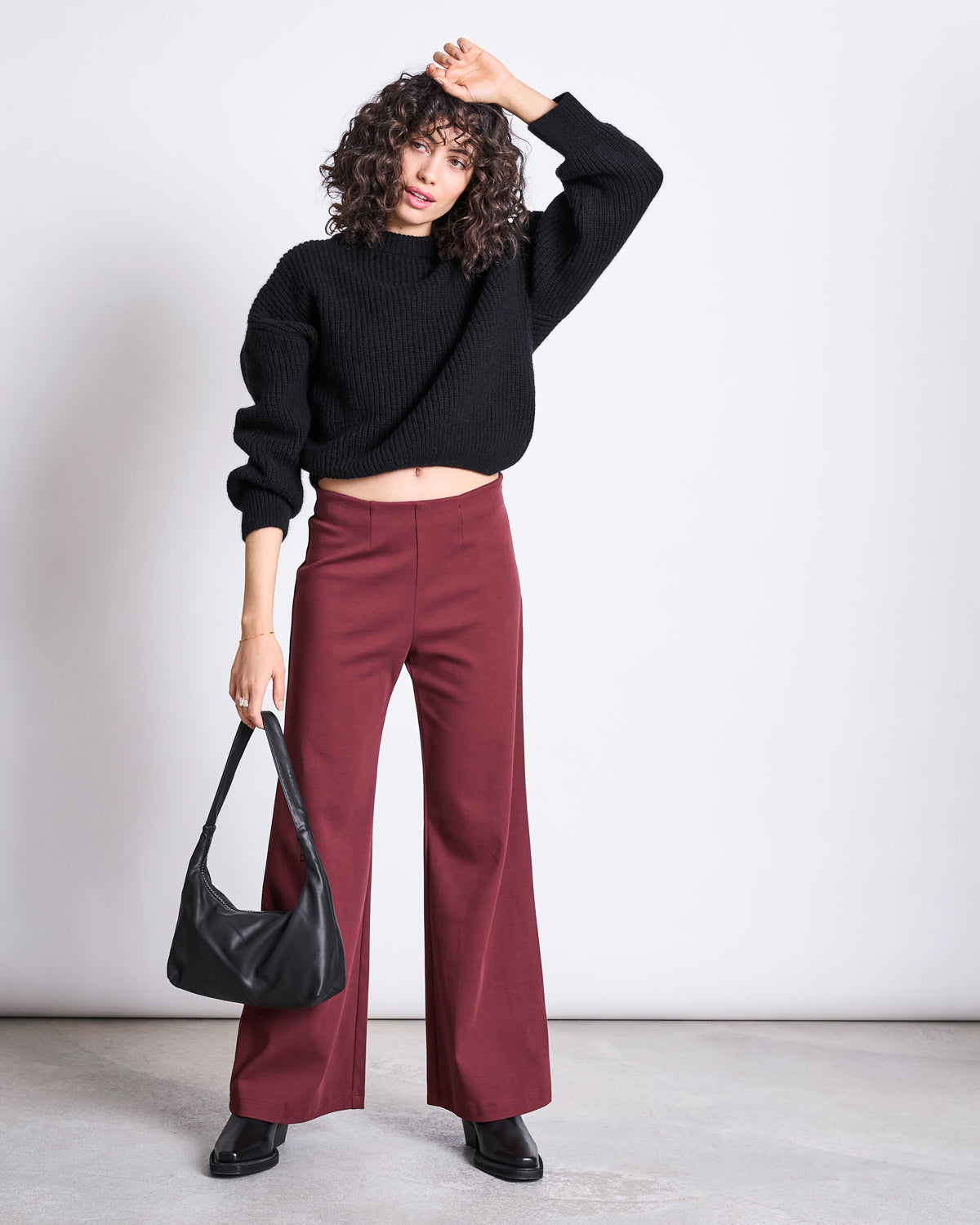 A woman poses against a plain background, wearing jan-n-junes WIDE PANTS KOHRA INTENSE RUST GOTS with a black knit sweater, black shoes, and a black handbag, raising one hand to her head.