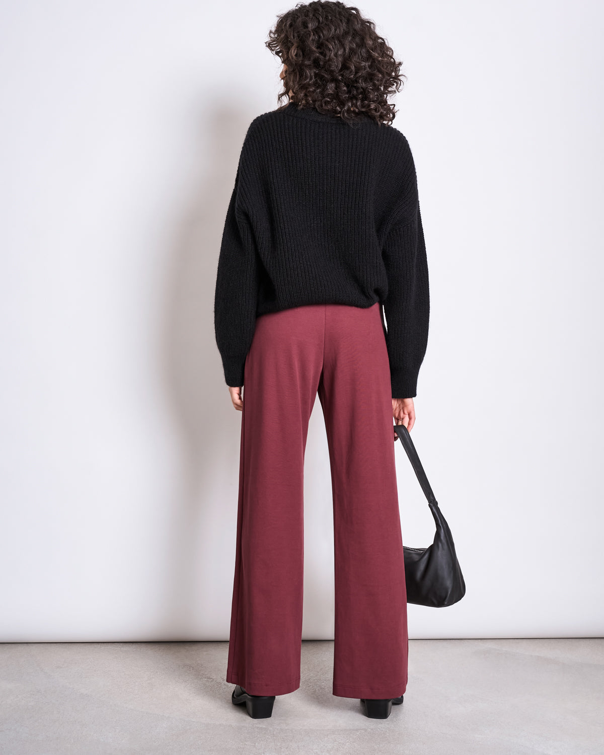 A person with curly hair faces away, wearing a black sweater, black heeled shoes, and holding a black handbag. Theyre dressed in jan-n-june’s WIDE PANTS KOHRA INTENSE RUST GOTS. The background is plain and light-colored.