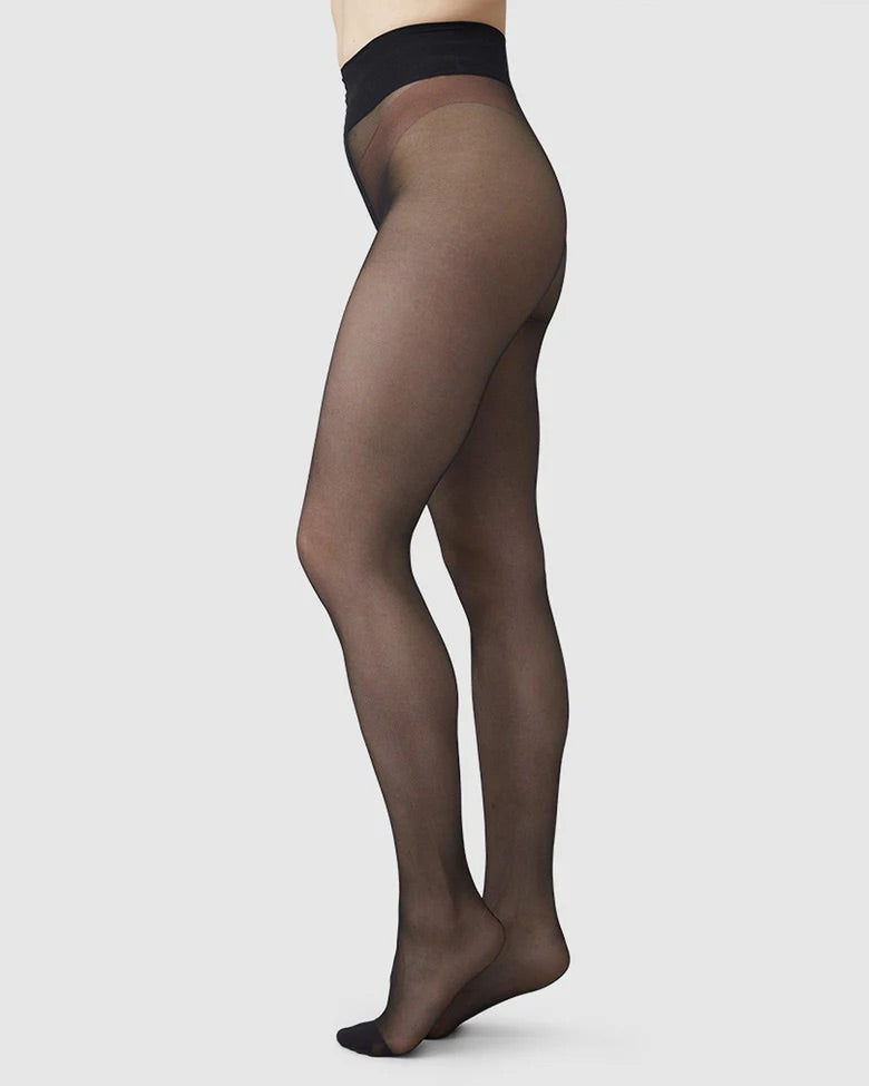 A person in profile wears jan-n-june SWEDISH STOCKINGS LADDER RESISTANT TIGHTS MALVA BLACK, made from recycled polyamide, showcasing their high waistband and semi-sheer fit against a plain light background.
