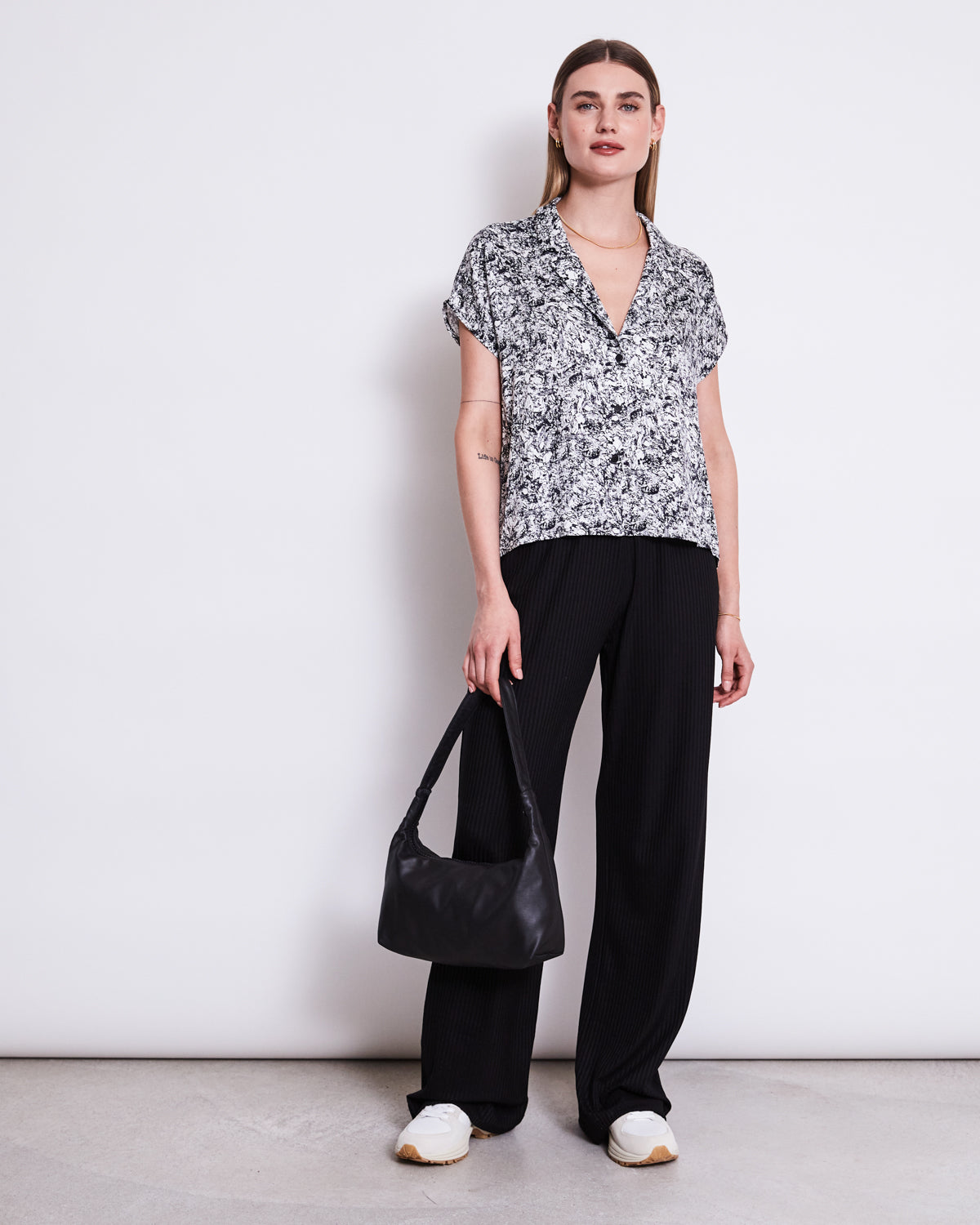 A woman with straight hair and a neutral expression stands against a white wall wearing the jan-n-june BLOUSE TIERRA NERVOUS PRINT BLACK, black wide-leg pants, white sneakers, and holding a black shoulder bag.