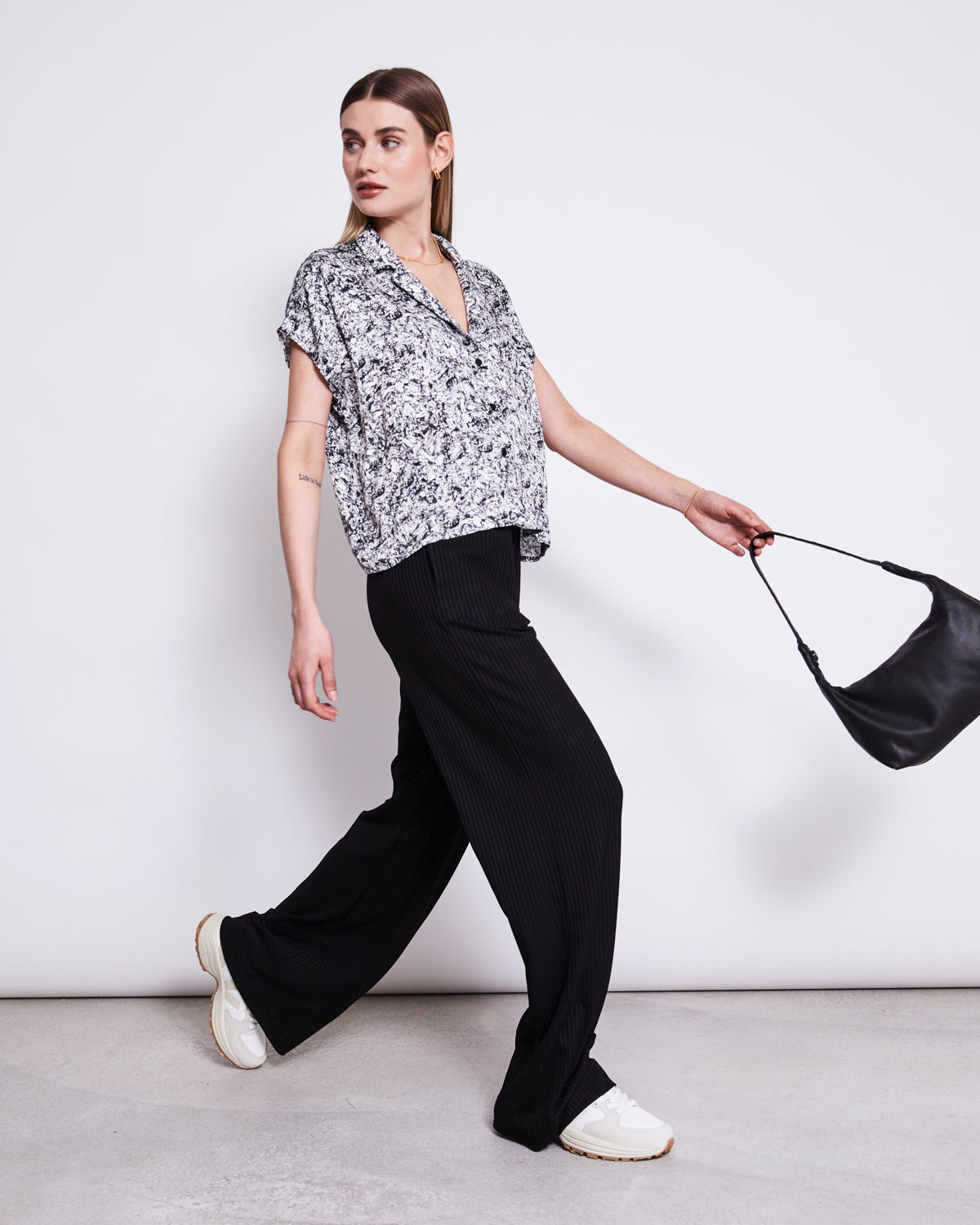 A woman with straight brown hair confidently wears the jan-n-june BLOUSE TIERRA NERVOUS PRINT BLACK, paired with black wide-leg pants and white sneakers, holding a small black handbag against a plain white background.