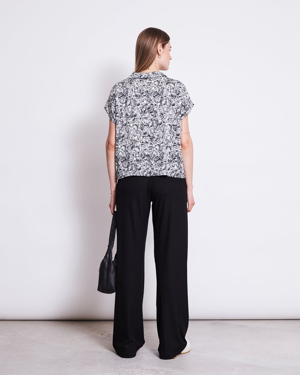 A woman with straight brown hair stands facing away, wearing the jan-n-june BLOUSE TIERRA NERVOUS PRINT BLACK with short sleeves and V-neckline, loose black pants, white shoes, and holding a black handbag against a plain white background.