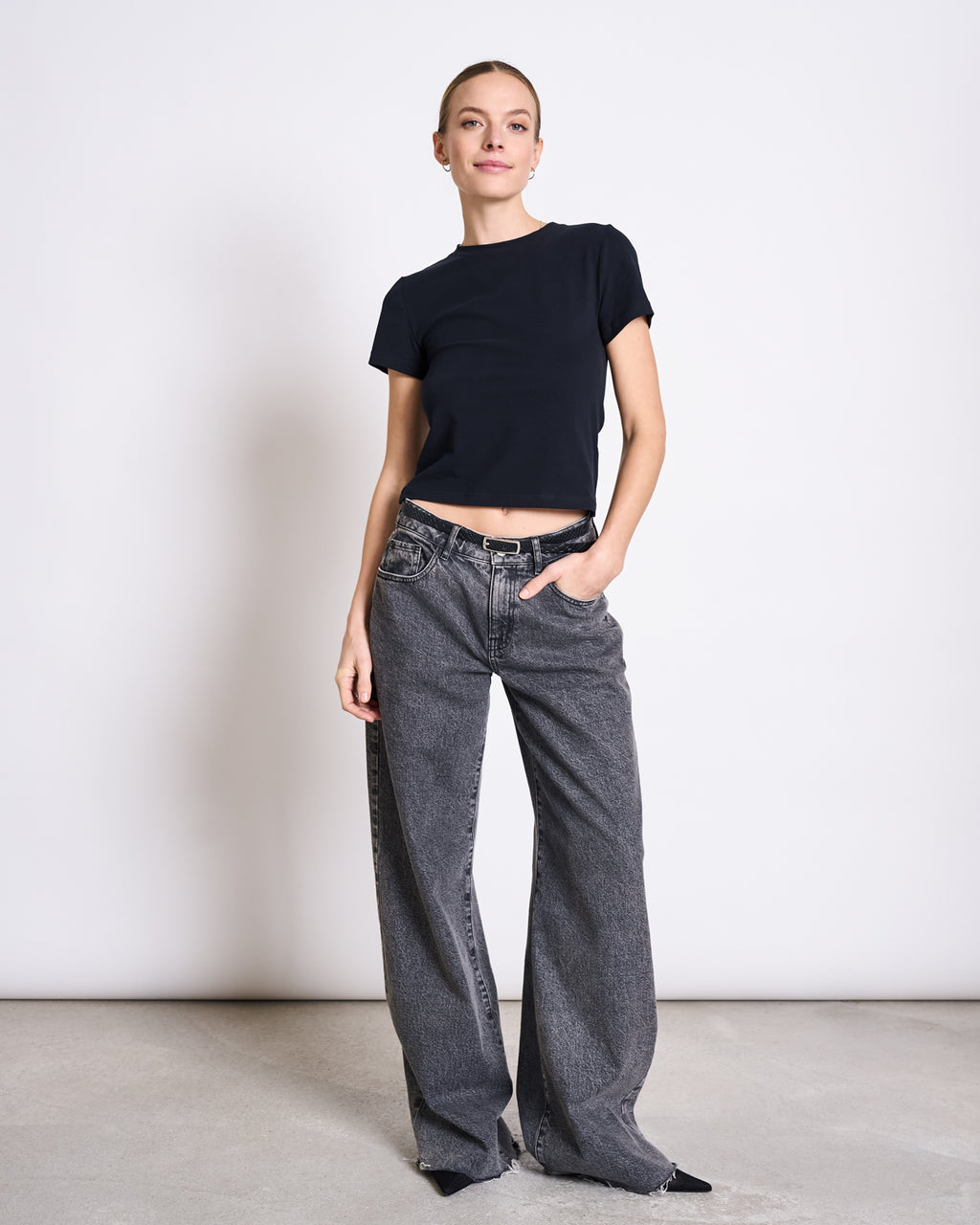 A woman poses against a white background in the jan-n-june T-SHIRT PALOOMA BLACK GOTS, paired with loose gray jeans and black pointed shoes. Her hair is tied back, one hand is in her pocket, and she smiles slightly.