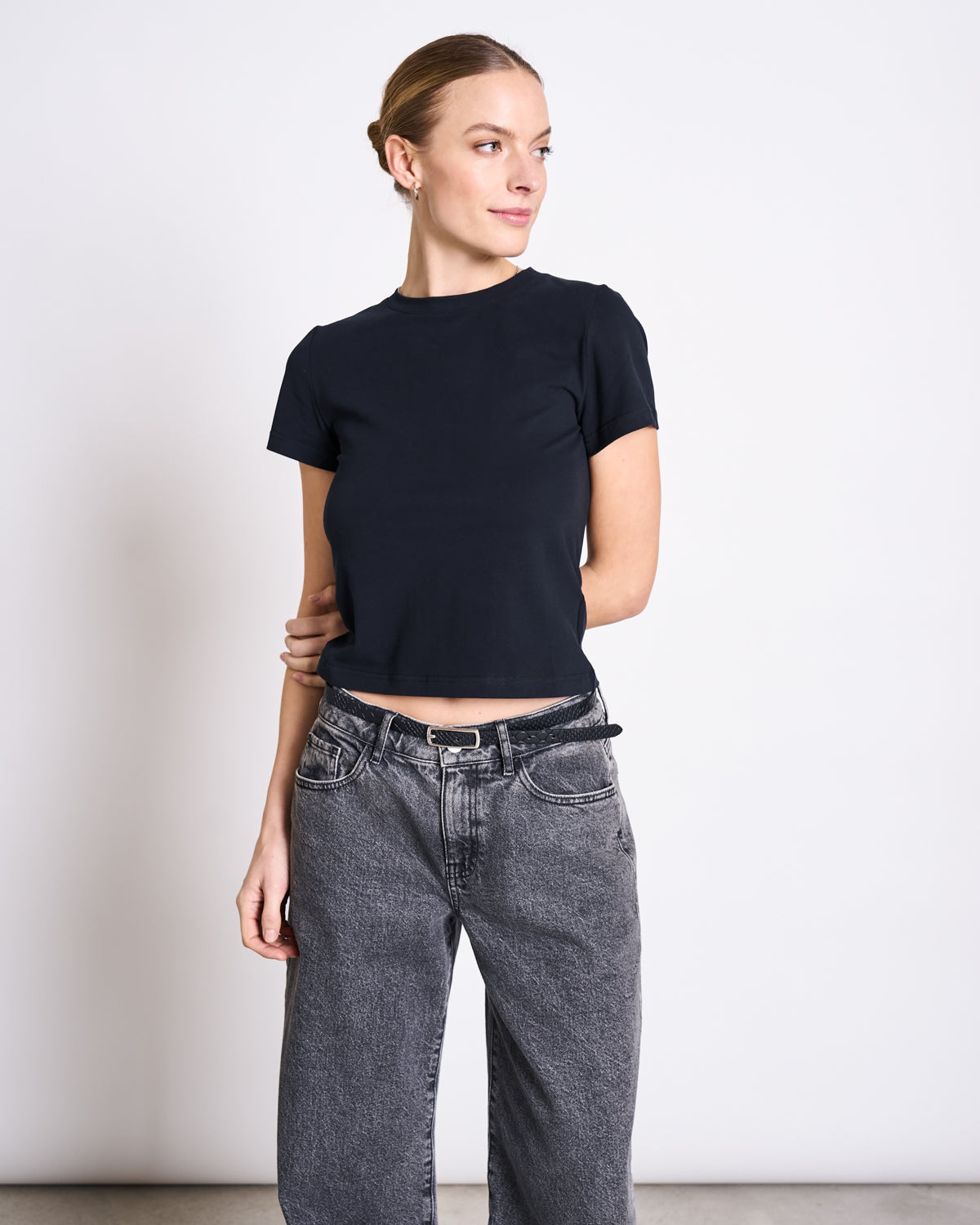 A woman with light hair in a bun wears the fitted jan-n-june T-SHIRT PALOOMA BLACK GOTS and loose gray jeans, standing against a plain white background and looking to her left with a slight smile.