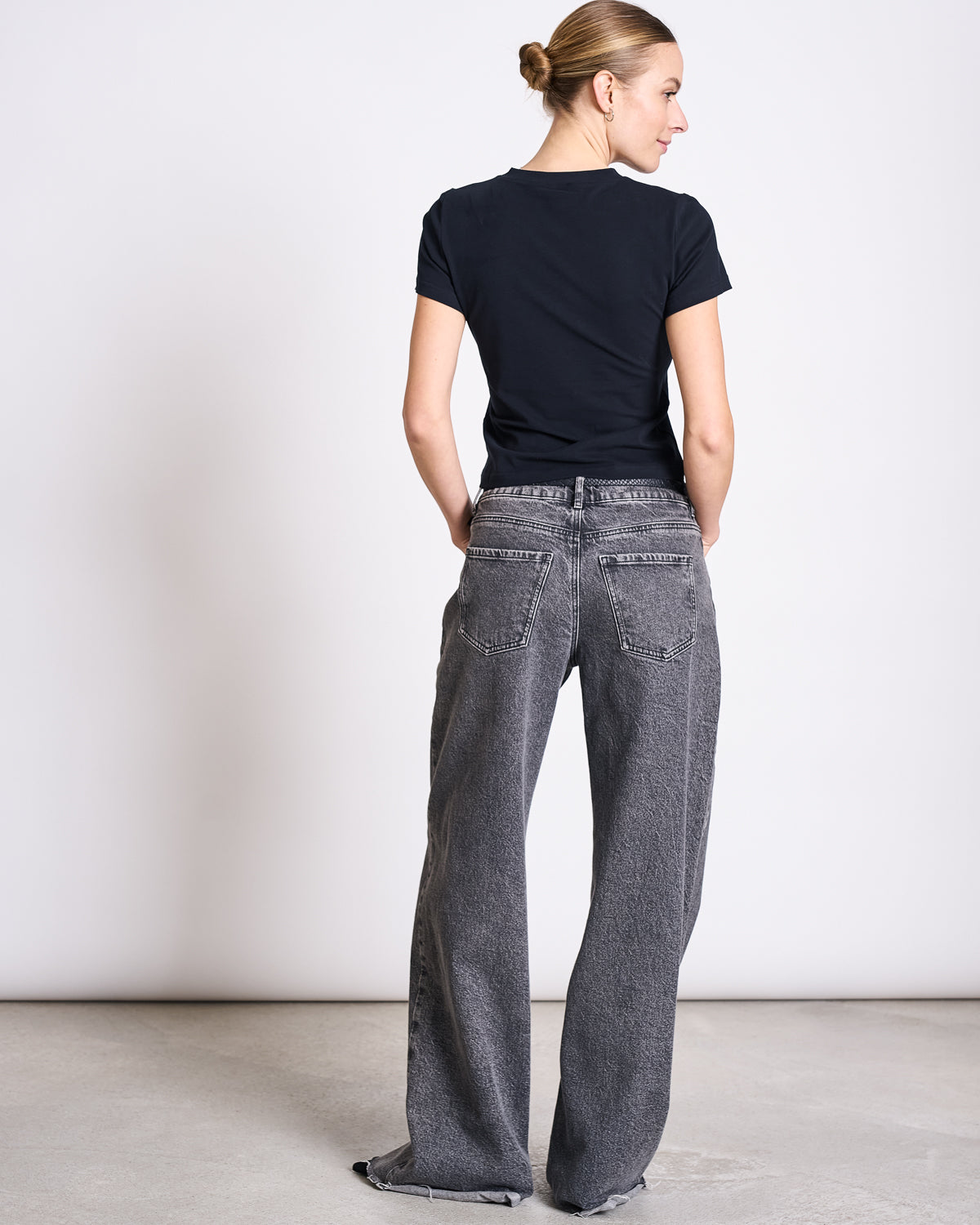 A woman with her hair in a neat bun faces away, wearing the jan-n-june T-SHIRT PALOOMA BLACK GOTS made from organic cotton and loose, faded gray jeans with frayed hems against a plain light background.