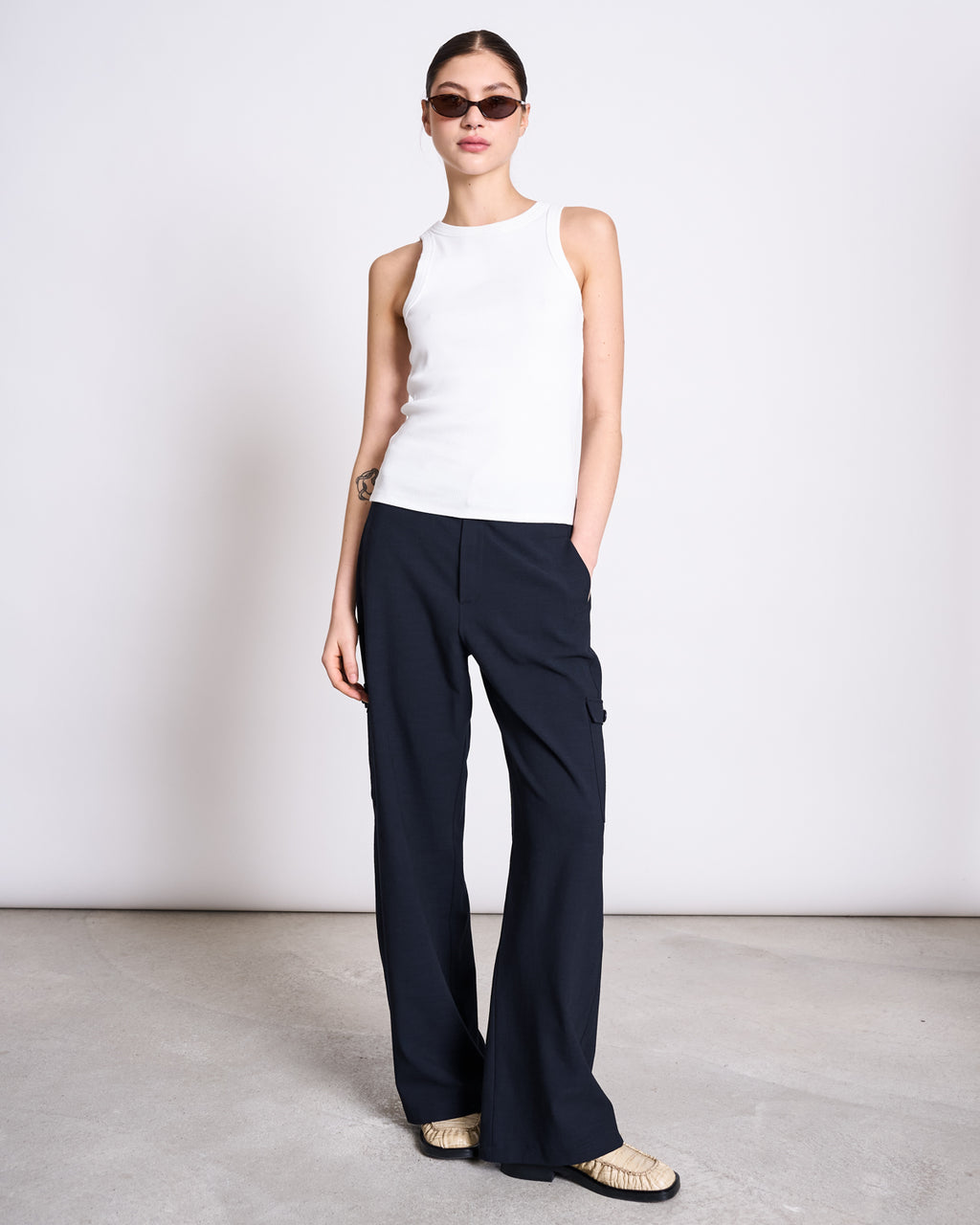 A woman in sunglasses and loose black pants stands against a plain light background wearing the jan-n-june ORGANIC COTTON TANK TOP PORTO LONG WHITE GOTS, one hand in her pocket.
