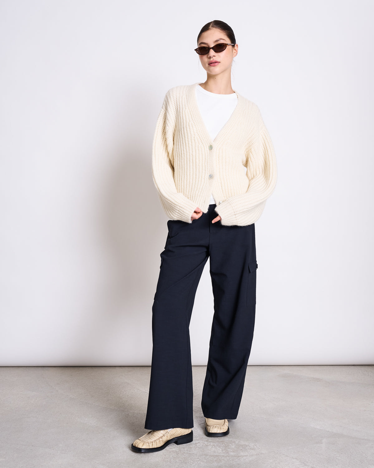 A woman with her hair pulled back, one hand in her pocket, wears sunglasses and the jan-n-june ORGANIC COTTON TANK TOP PORTO LONG WHITE GOTS under a white knit cardigan, with slim dark wide-leg pants and beige snakeskin-patterned shoes.