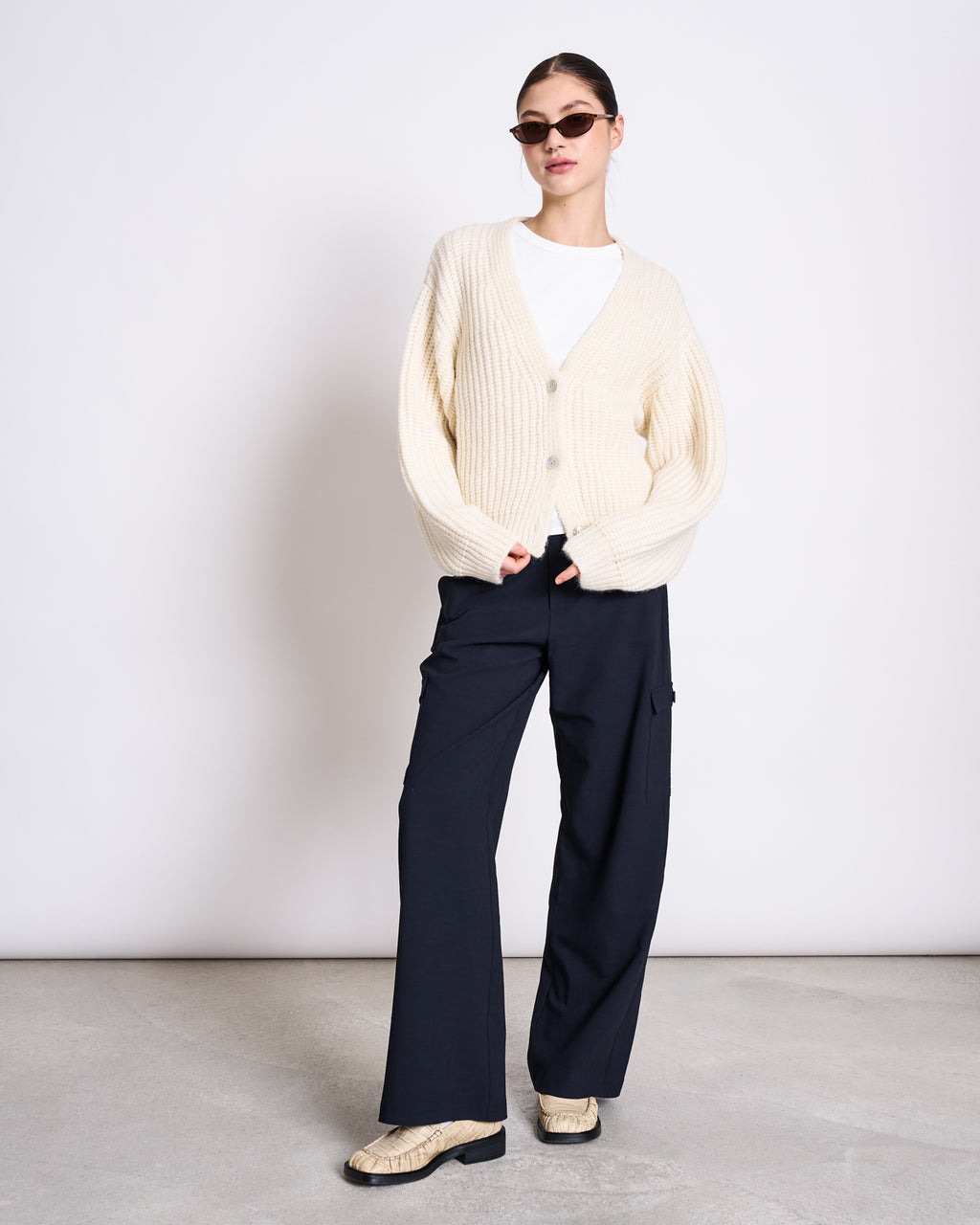 A woman stands against a plain background wearing sunglasses, a cream chunky knit cardigan over a white top, jan-n-june CARGO PANTS FANO NIGHTFALL, and snakeskin-patterned loafers.