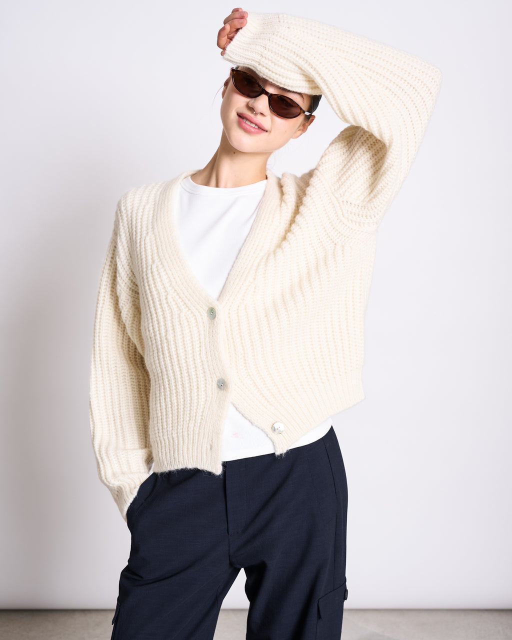 A person wearing jan-n-june’s CARGO PANTS FANO NIGHTFALL, sunglasses, a cream knit cardigan over a white top poses with one arm raised and bent overhead, smiling against a plain background.