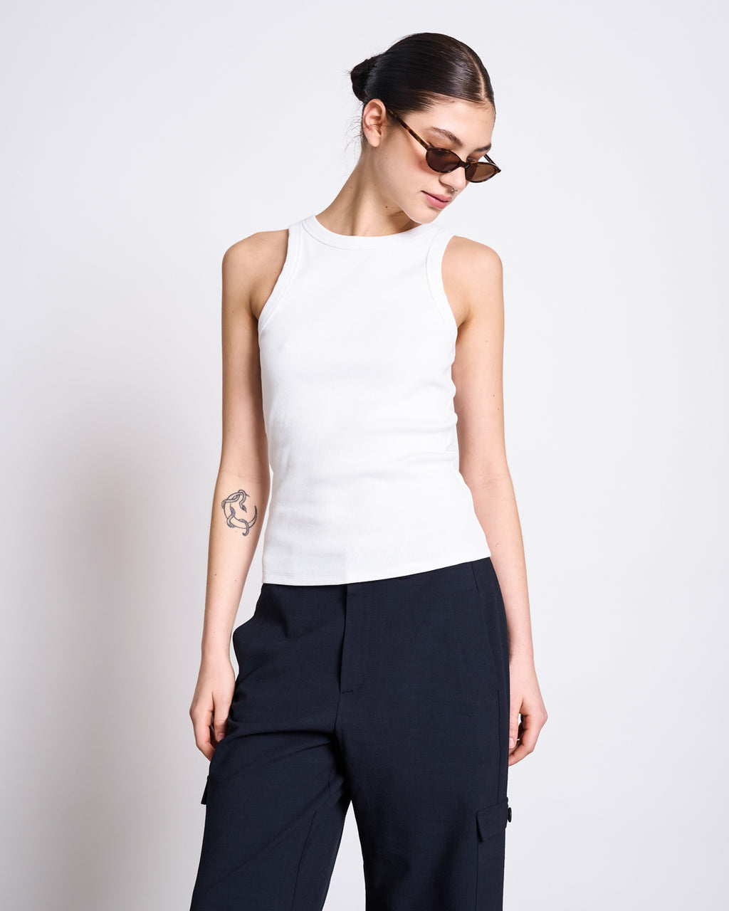 A woman in the jan-n-june ORGANIC COTTON TANK TOP PORTO LONG WHITE GOTS, styled with dark pants and sunglasses, stands against a plain background. Her look, complete with a low bun and forearm tattoo, is perfect for everyday comfort.