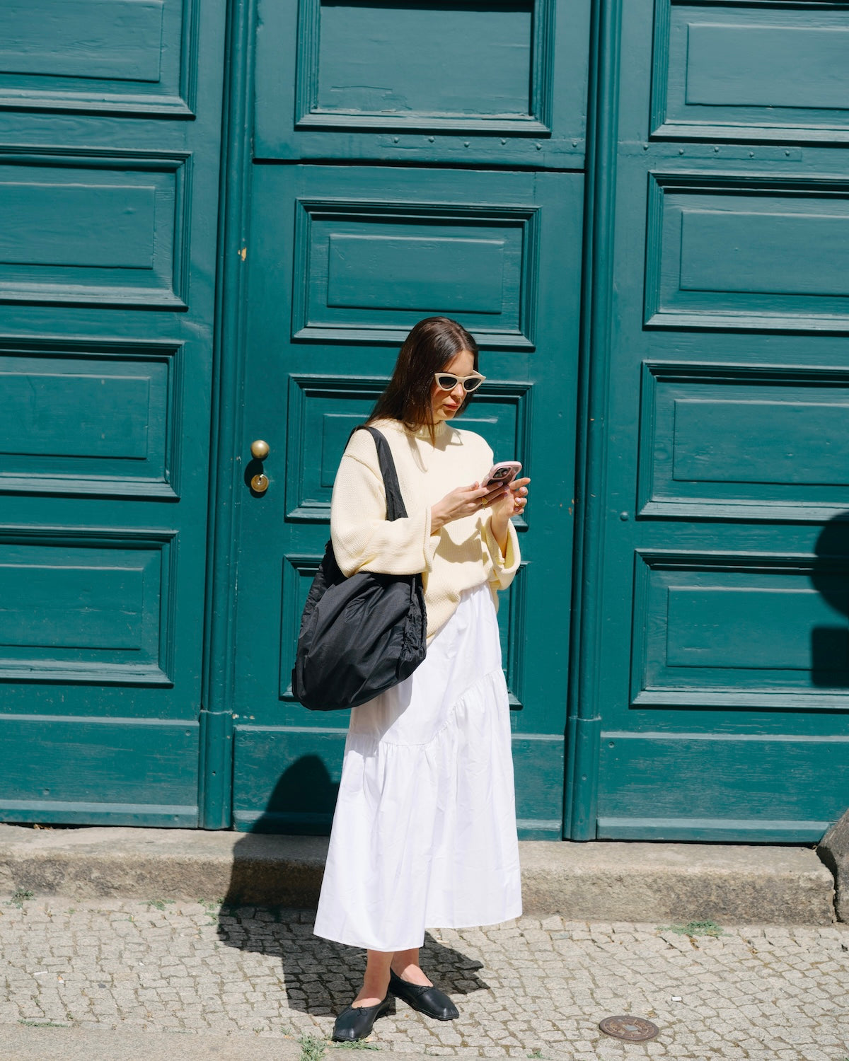 A woman stands on a cobblestone sidewalk in front of a large teal door, looking at her phone. She wears the jan-n-june JUMPER SONA BUTTERMILK GOTS organic cotton sweater and carries a large black bag on her shoulder.
