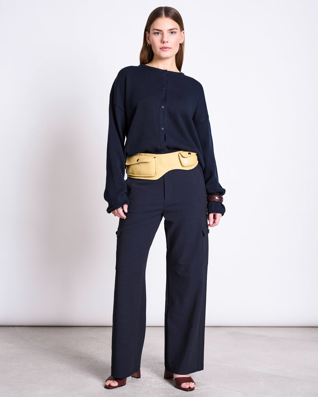 A woman faces forward in a loose navy button-up, matching wide-leg pants, yellow belt bag, and open-toe heels. She layers with jan-n-june's LIGHT CARDIGAN ASAS NIGHTFALL GOTS against a plain white background.