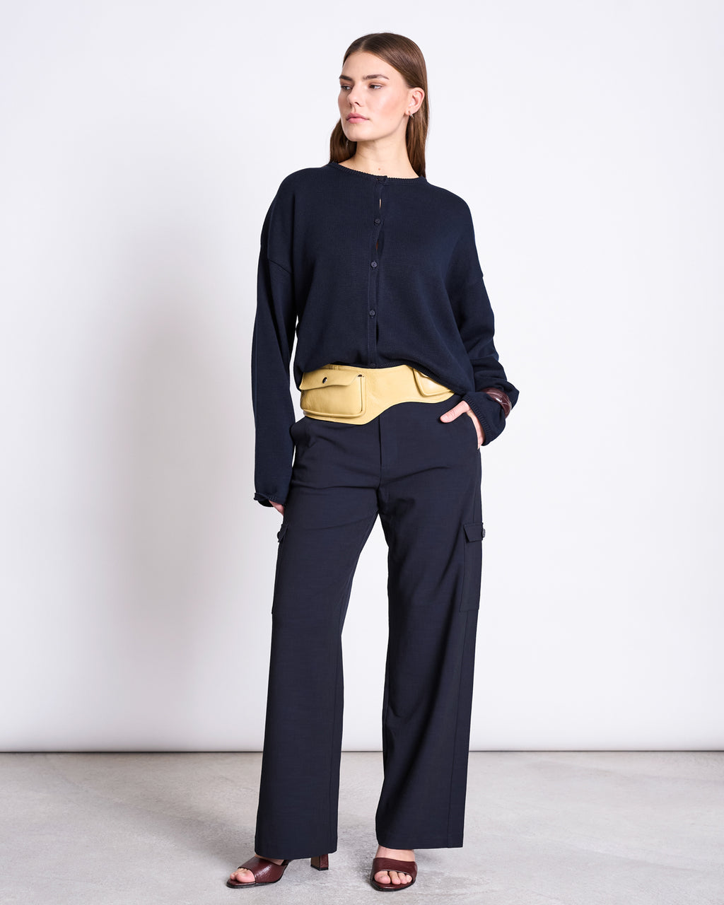 A woman stands against a plain background wearing the jan-n-june LIGHT CARDIGAN ASAS NIGHTFALL GOTS with matching wide-leg pants, maroon open-toe heels, and a beige belt bag. She looks to the side with a neutral expression.