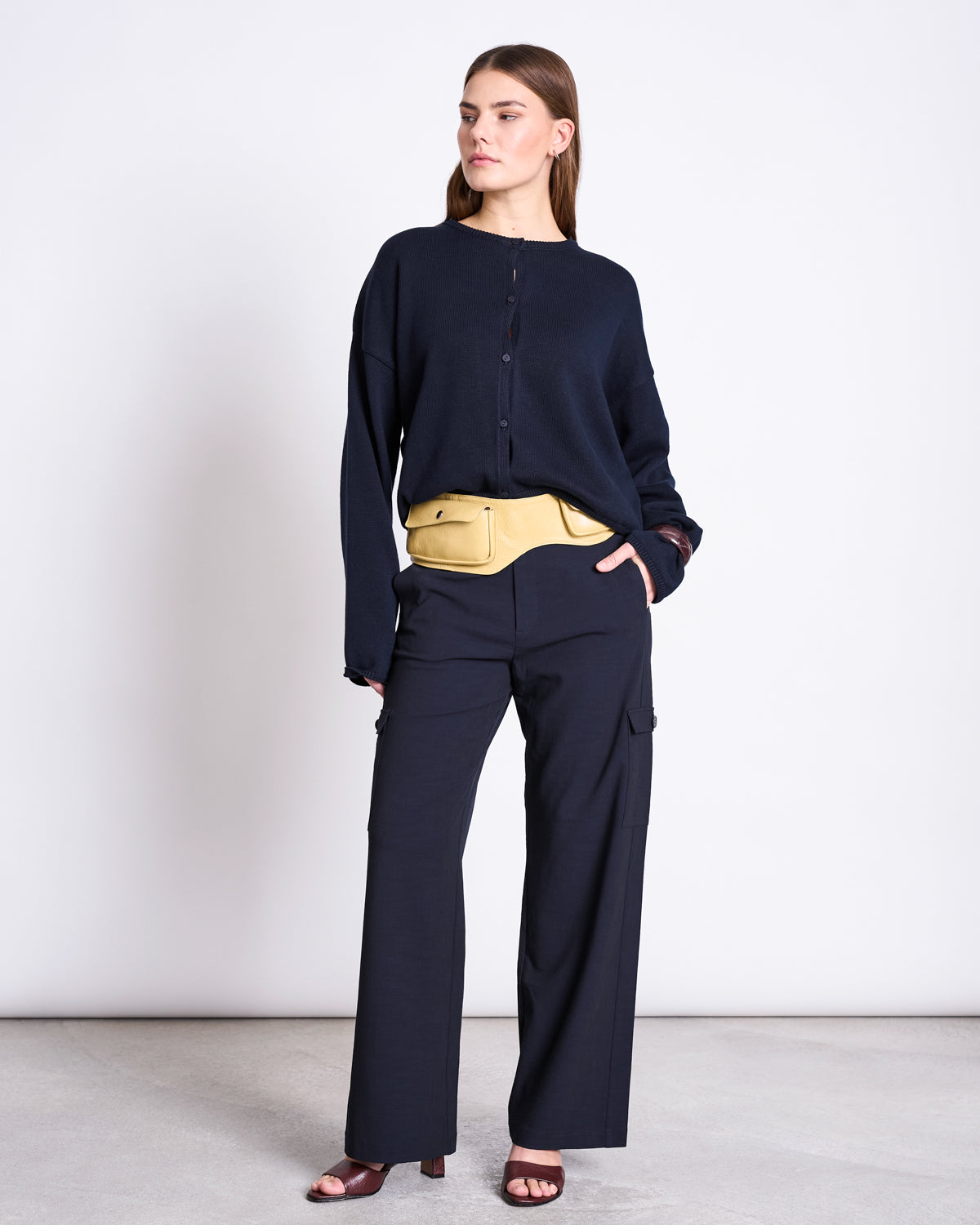 A woman stands against a plain background wearing the jan-n-june LIGHT CARDIGAN ASAS NIGHTFALL GOTS with matching wide-leg pants, maroon open-toe heels, and a beige belt bag. She looks to the side with a neutral expression.