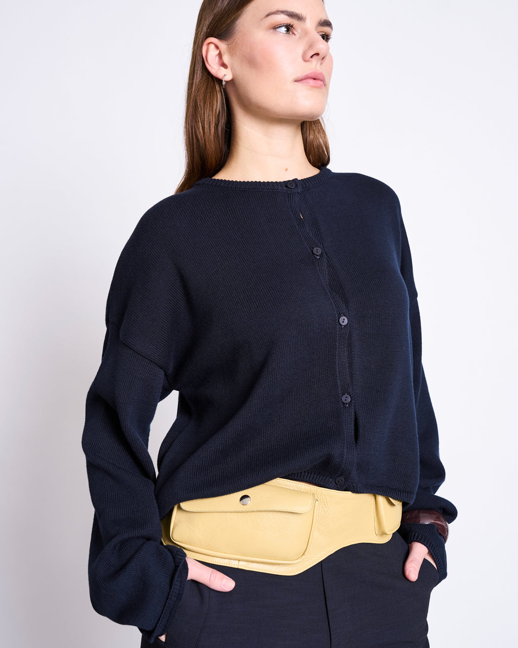 A woman stands confidently against a light background wearing the jan-n-june LIGHT CARDIGAN ASAS NIGHTFALL GOTS in dark navy, paired with black pants and a light yellow belt bag around her waist.