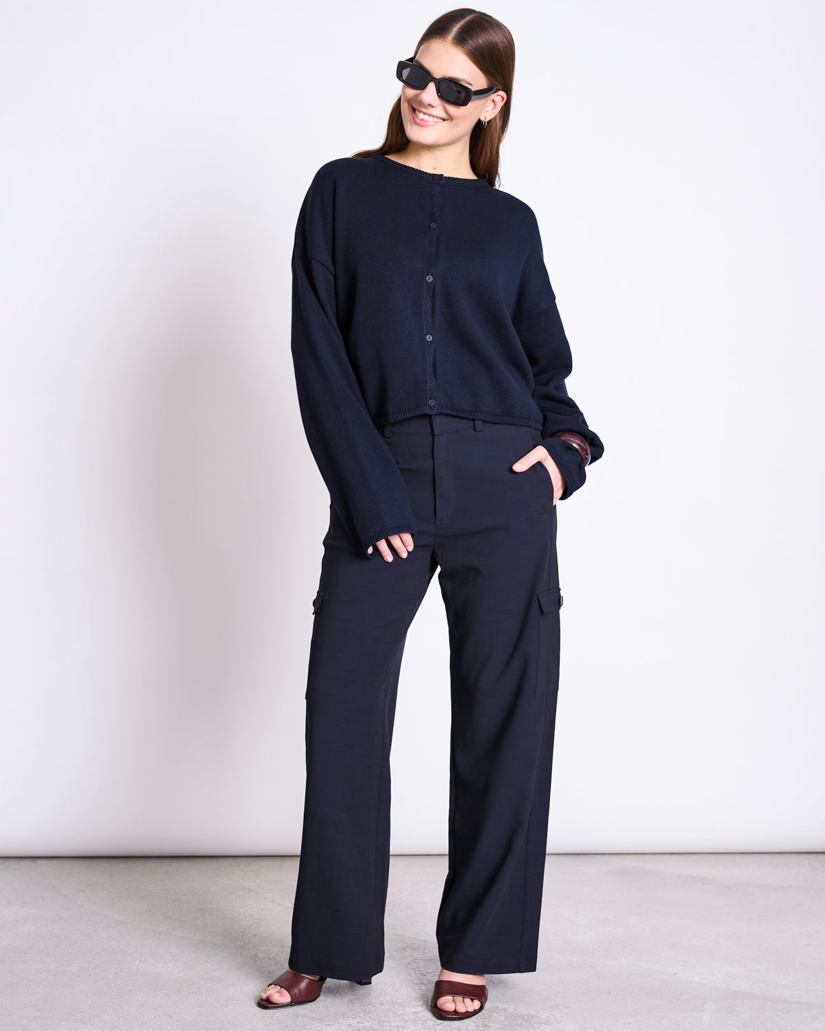 A woman smiles against a plain background, wearing jan-n-june's LIGHT CARDIGAN ASAS NIGHTFALL GOTS over a dark sweater and wide-leg pants, paired with sunglasses and open-toe heels. One hand is in her pocket, the other relaxed by her side.