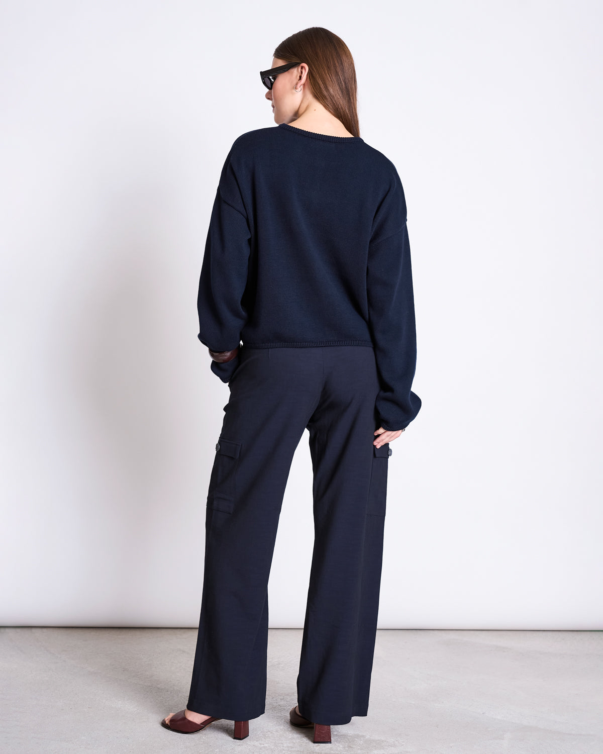 A woman with long brown hair, facing away against a white background, wears jan-n-june's LIGHT CARDIGAN ASAS NIGHTFALL GOTS—an organic cotton black cardigan—with wide-leg pants, brown heeled shoes, and sunglasses.