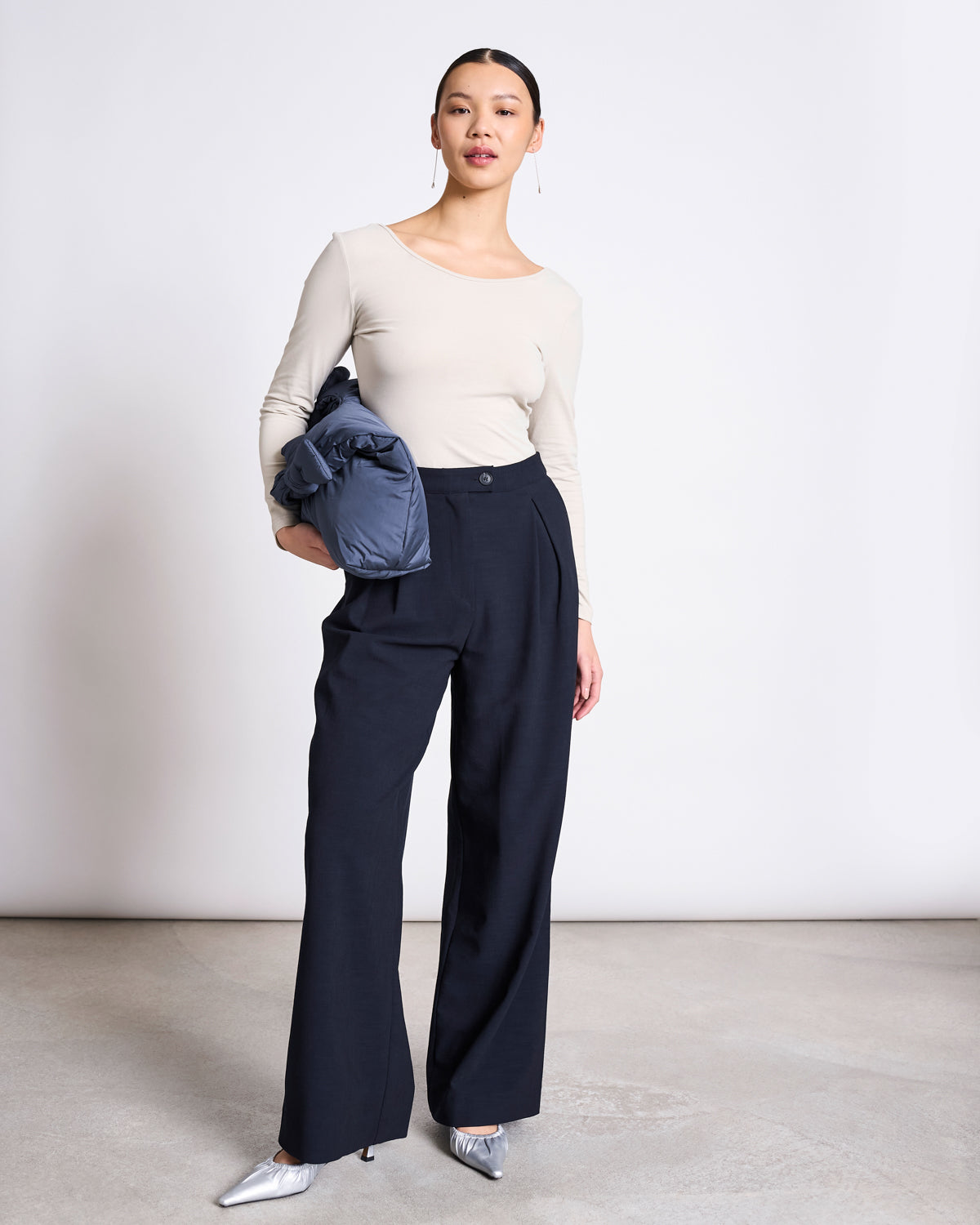 A woman stands against a plain background wearing the jan-n-june BALLERINA LONGSLEEVE LEON IVORY GOTS top in organic cotton, paired with high-waisted navy wide-leg trousers, silver heels, and holding a folded navy jacket.