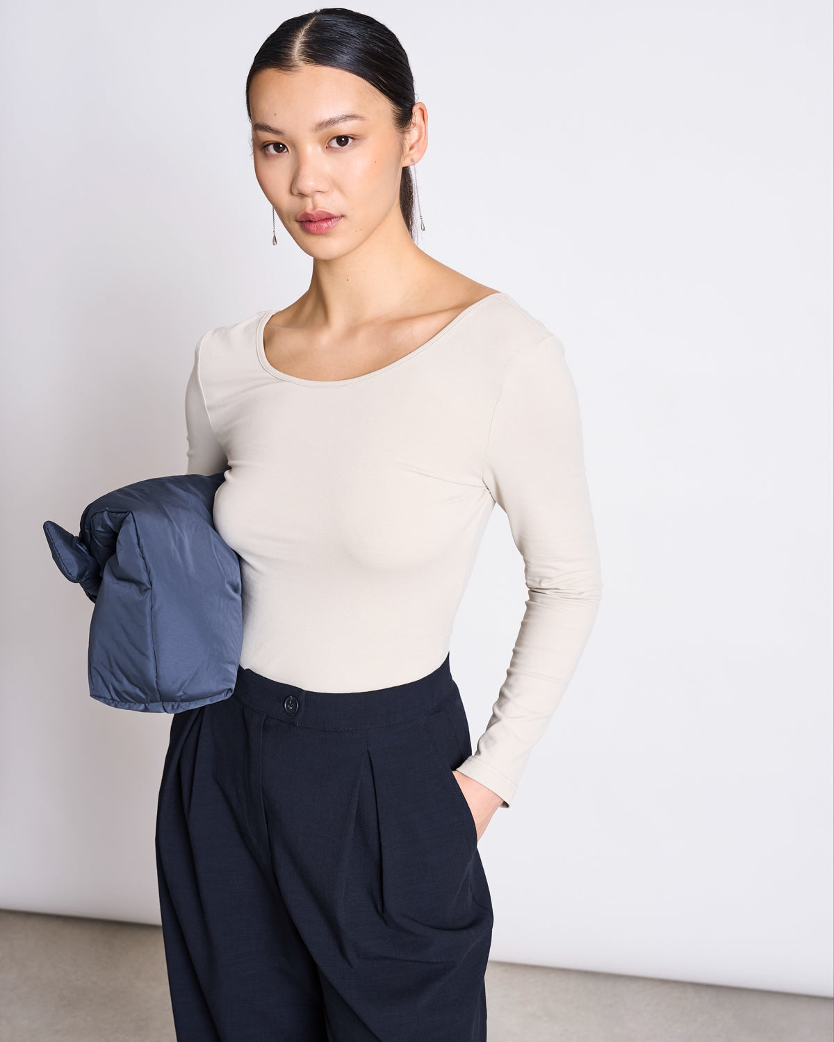 A woman with slicked-back hair wears the jan-n-june BALLERINA LONGSLEEVE LEON IVORY GOTS top and high-waisted black trousers, standing with one hand in her pocket and holding a blue jacket against a plain light background.