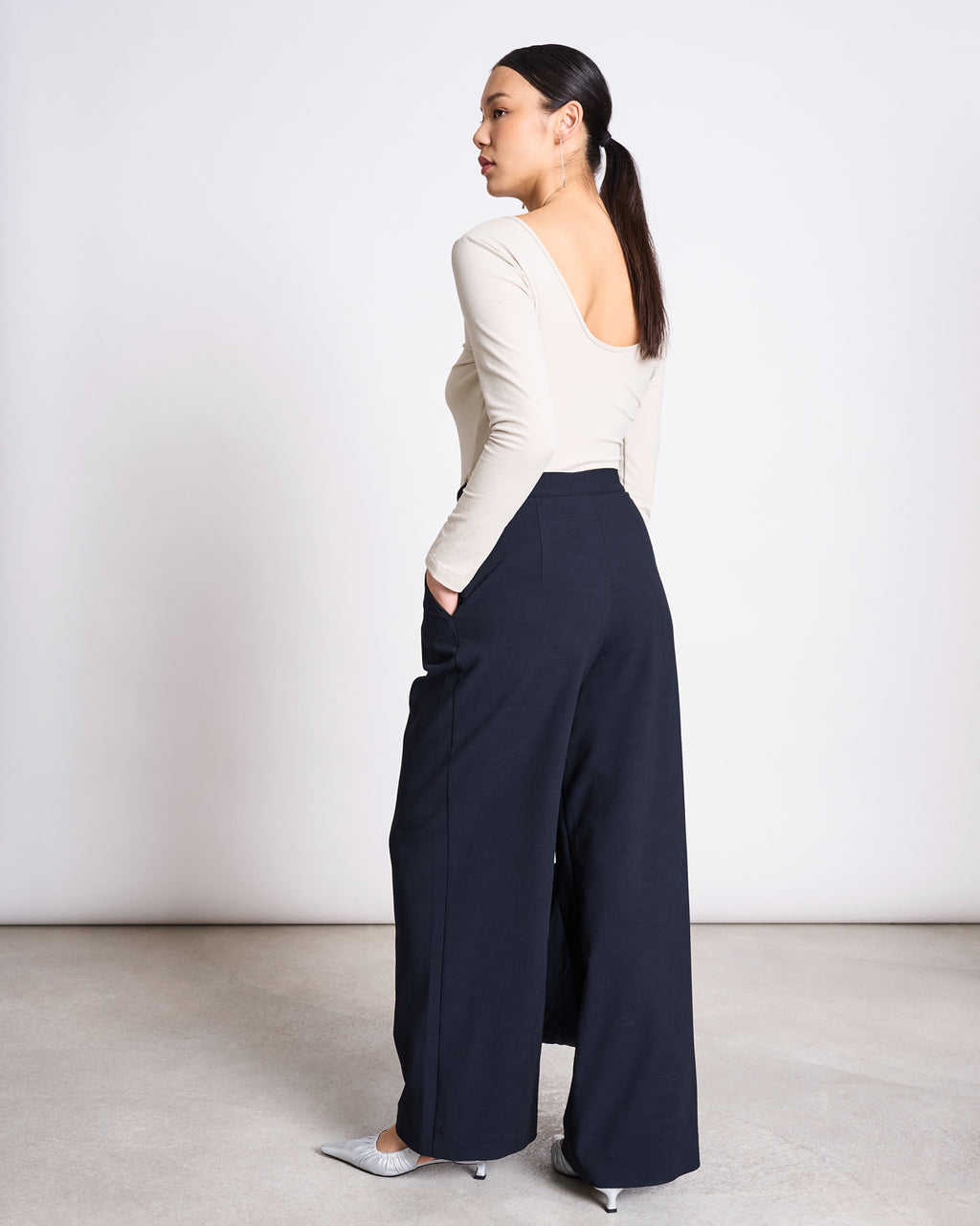 A woman with long dark hair in a low ponytail wears the jan-n-june BALLERINA LONGSLEEVE LEON IVORY GOTS top, wide-leg black pants, and pointed white heels. She stands hands-in-pockets, looking to the side against a plain background.