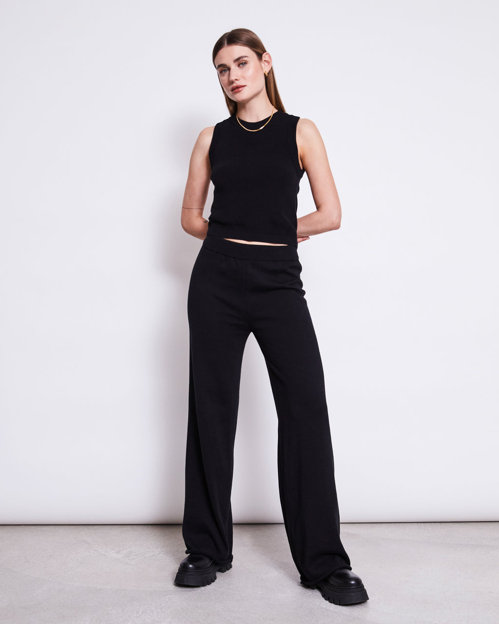 A woman with straight brown hair poses against a white wall, wearing jan-n-junes KNIT PANTS ANA BLACK GOTS with an elastic waistband, a sleeveless black top, and black platform shoes, her hands placed behind her back.