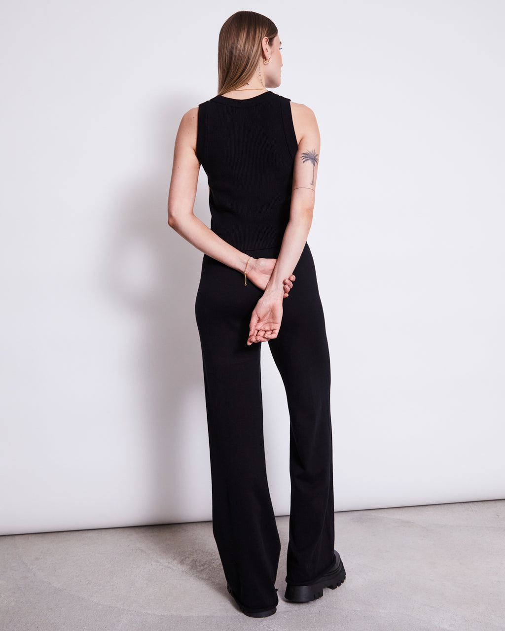 A woman stands with her back to the camera, wearing jan-n-june KNIT PANTS ANA BLACK GOTS—organic cotton pants with an elastic waistband—paired with a sleeveless black top, black shoes, and a palm tree tattoo on her upper left arm.
