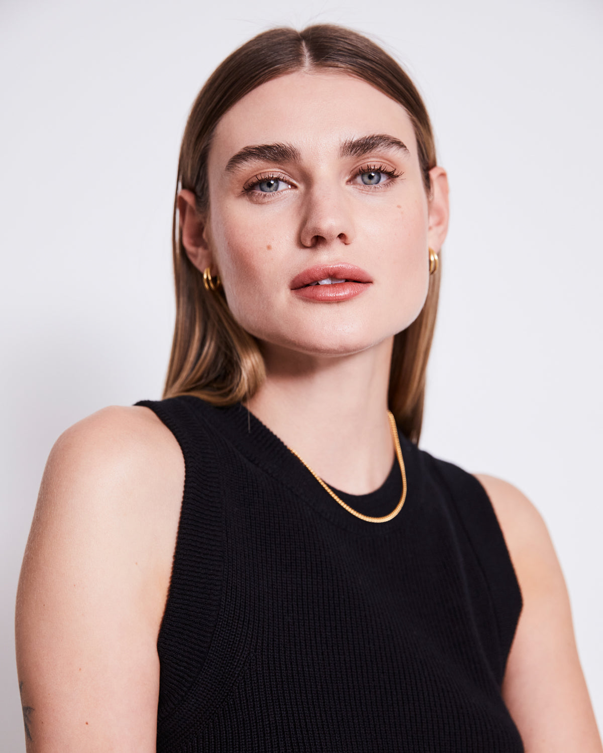 A woman with straight light brown hair and blue eyes wears a sleeveless black top, gold jewelry, and jan-n-june’s KNIT PANTS ANA BLACK GOTS with an elastic waistband, posing confidently against a plain white background.