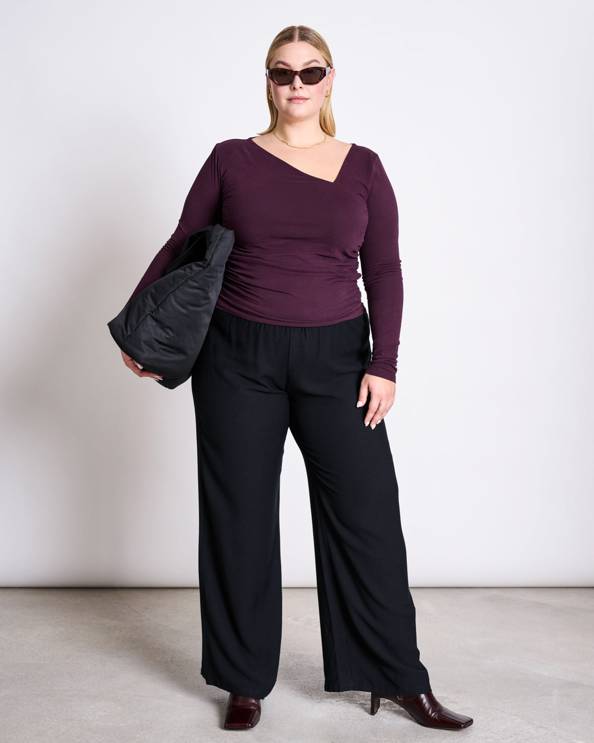 A woman wearing the jan-n-june ASYMMETRICAL LONGSLEEVE CASMA CHERRY LACQUER GOTS top, black wide-leg pants, burgundy boots, and sunglasses stands against a plain background, holding a black bag under her arm.