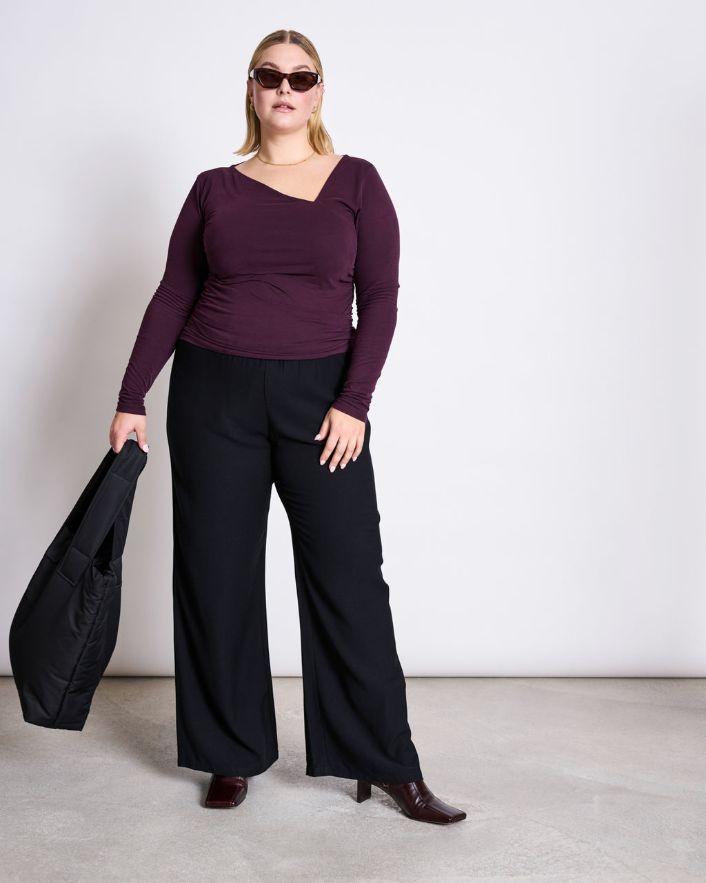 A woman in jan-n-junes ASYMMETRICAL LONGSLEEVE CASMA CHERRY LACQUER GOTS top, sunglasses, wide-leg black pants, heeled boots, and a black bag stands by a white wall.