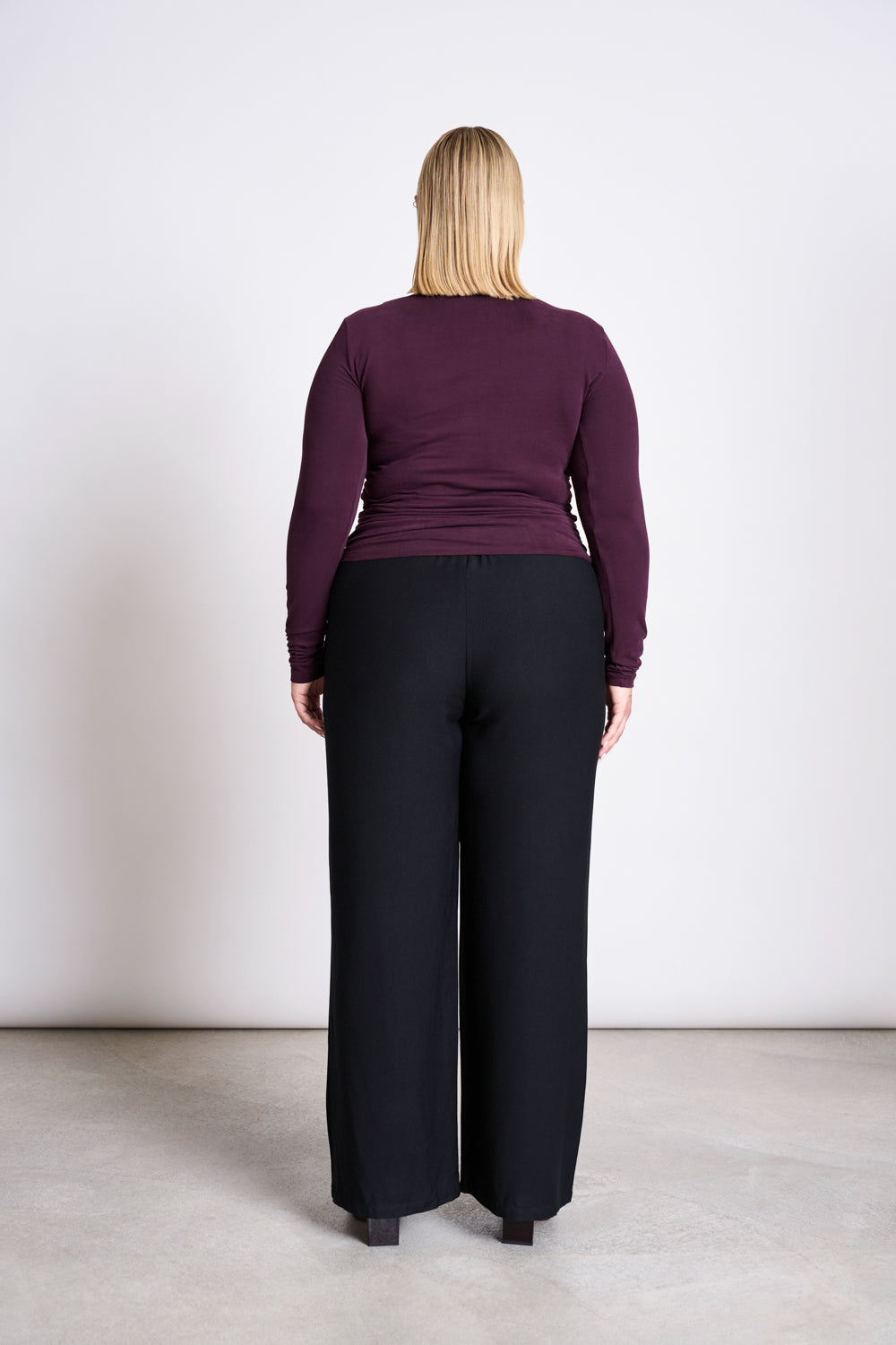 A person with straight blonde hair faces away from the camera, wearing the jan-n-june ASYMMETRICAL LONGSLEEVE CASMA CHERRY LACQUER GOTS top and loose black pants, against a plain white wall and light-colored floor.