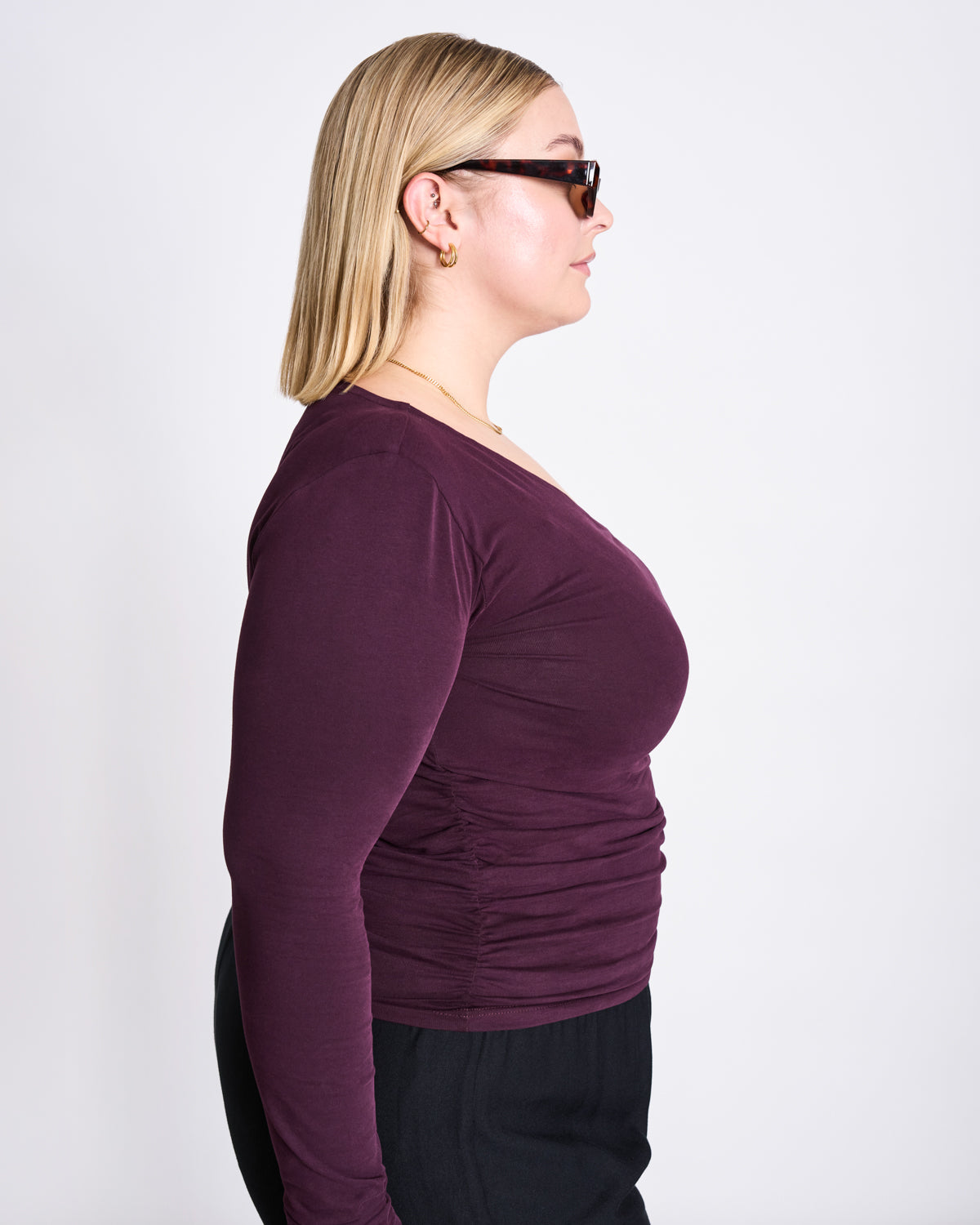 A blonde woman in dark sunglasses, gold hoop earrings, black pants, and the jan-n-june ASYMMETRICAL LONGSLEEVE CASMA CHERRY LACQUER GOTS top stands in profile against a plain white background.