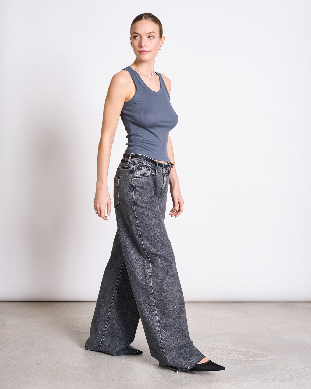 A woman with her hair tied back wears a fitted jan-n-june MINIRIB TANK TOP AMAGER TURBULENCE GOTS, wide-leg gray jeans, and black pointed-toe shoes, standing against a plain white background.