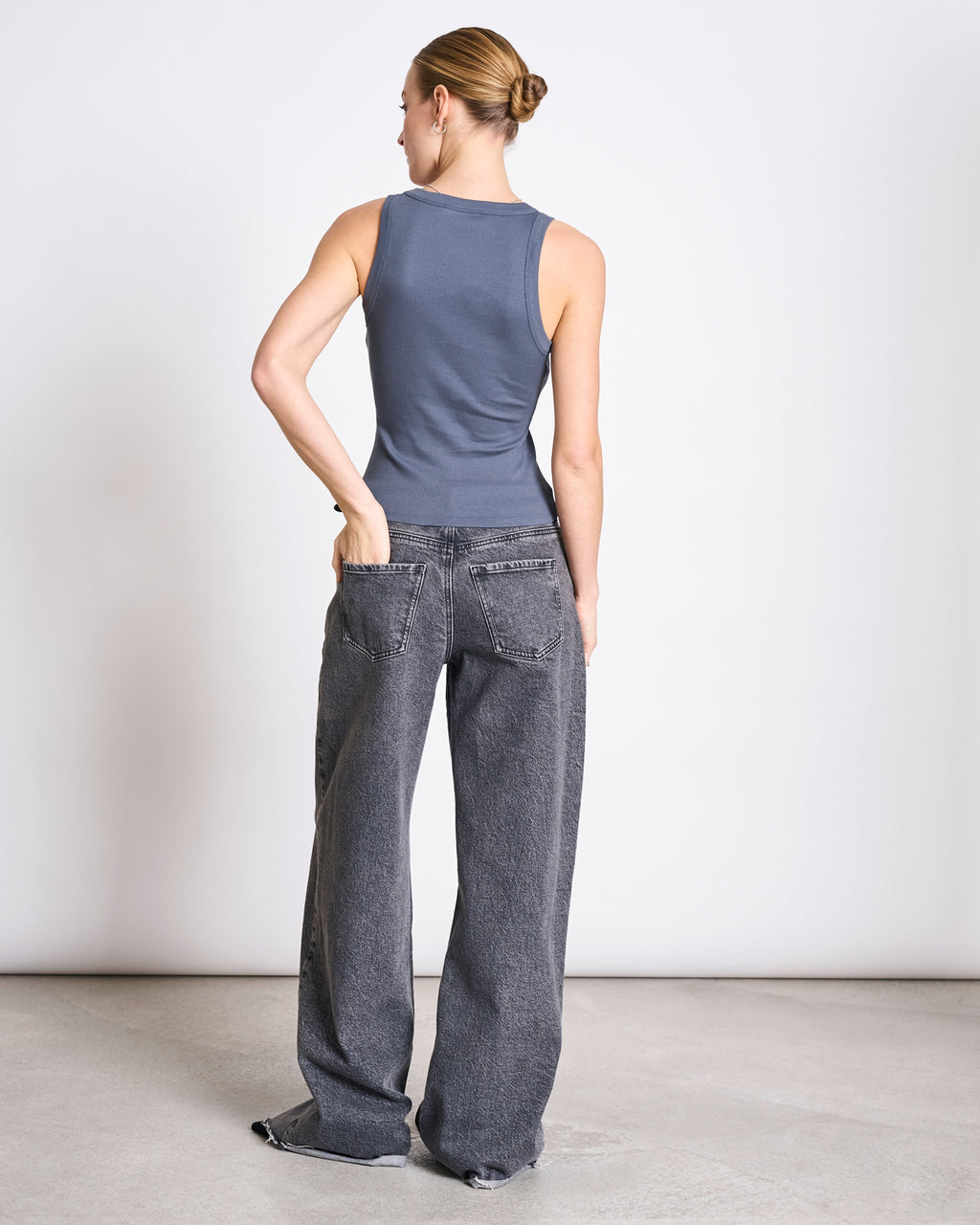 A woman with her hair in a bun stands on a light concrete floor, wearing the jan-n-june MINIRIB TANK TOP AMAGER TURBULENCE GOTS and loose, faded black jeans with frayed hems, one hand in her back pocket, facing away against a plain wall.