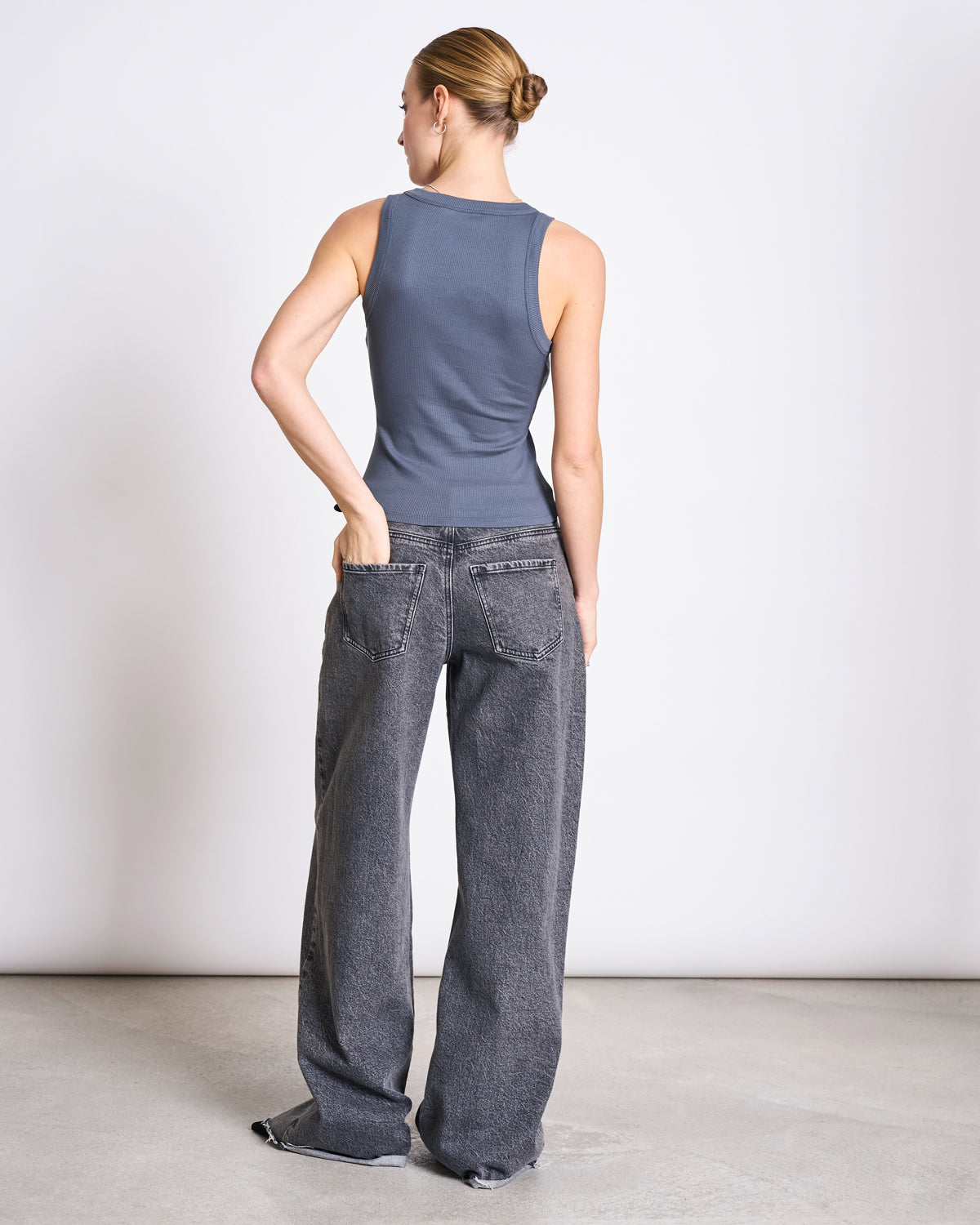 A woman with her hair in a bun stands on a light concrete floor, wearing the jan-n-june MINIRIB TANK TOP AMAGER TURBULENCE GOTS and loose, faded black jeans with frayed hems, one hand in her back pocket, facing away against a plain wall.