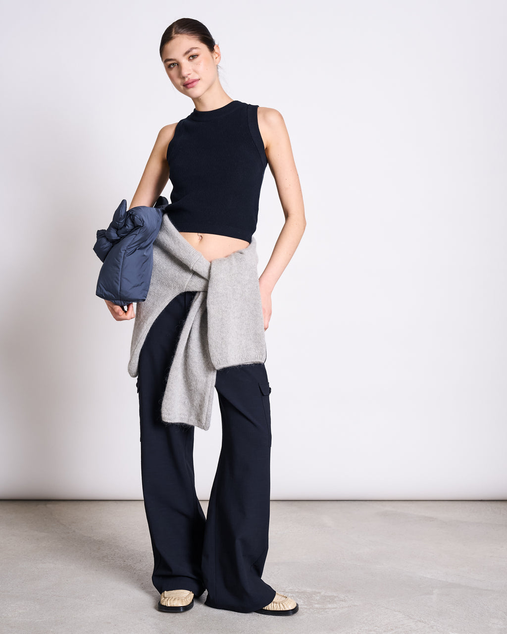 A woman stands against a plain background wearing the jan-n-june KNIT TOP GARDA NIGHTFALL GOTS, paired with black wide-leg pants, beige shoes, a gray sweater tied at her waist, and a blue jacket draped over her arm for an effortless look.
