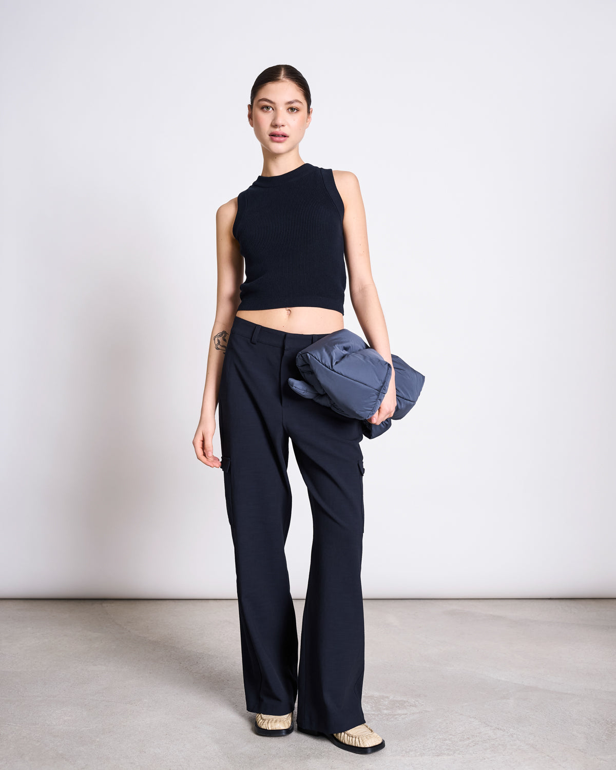 A woman with straight brown hair pulled back stands in a minimalist room wearing the jan-n-june KNIT TOP GARDA NIGHTFALL GOTS, navy blue trousers, and beige shoes, holding a folded blue jacket and showing a neutral expression.