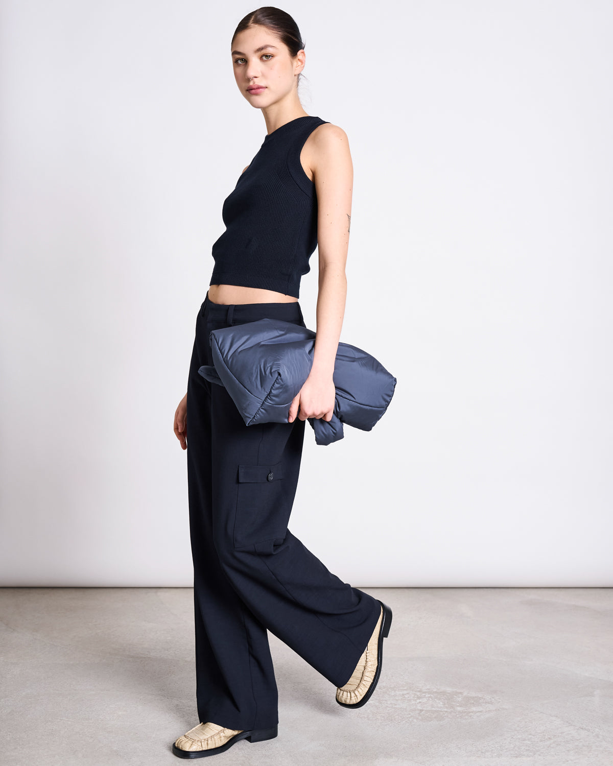 A woman stands in profile against a plain background, wearing the jan-n-june KNIT TOP GARDA NIGHTFALL GOTS, wide-leg black pants, and beige shoes. She holds a folded gray jacket under one arm and looks toward the camera.