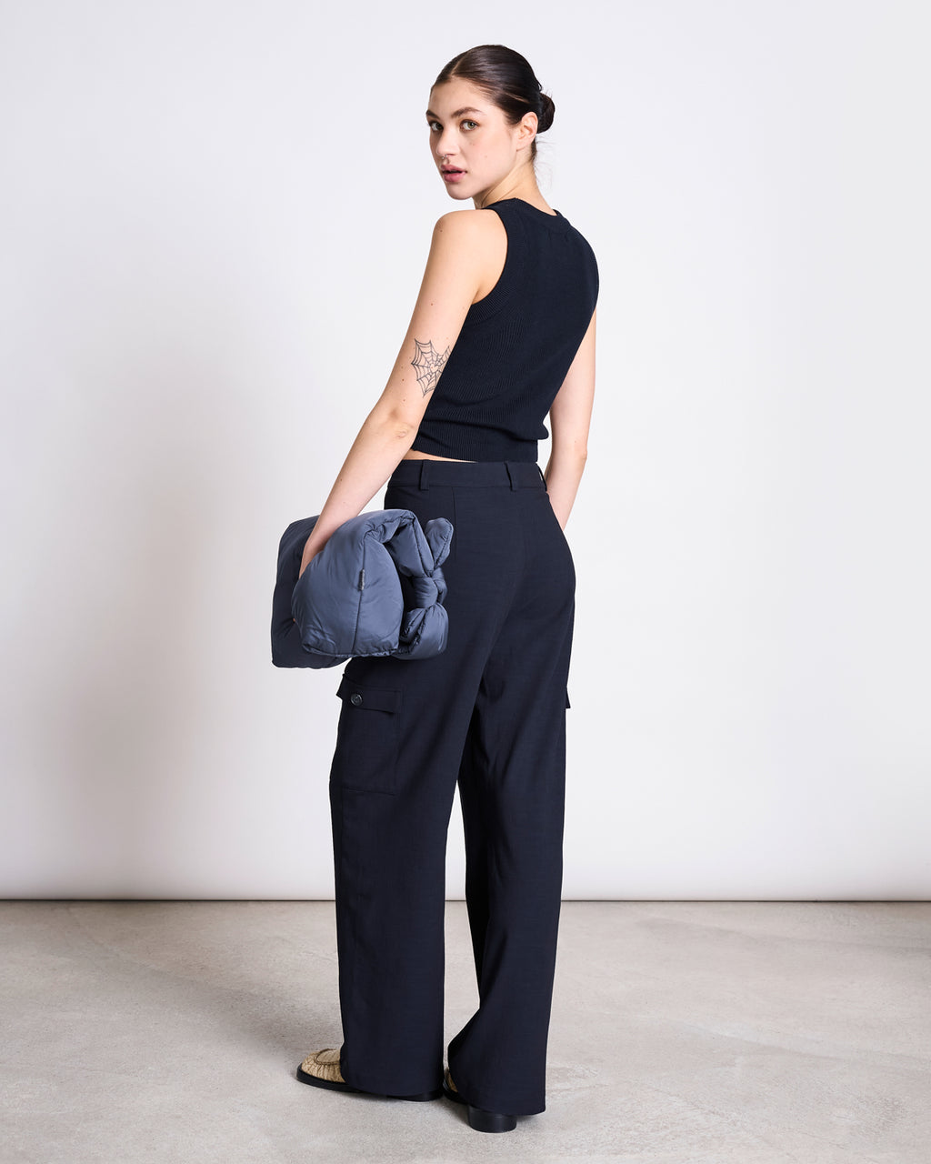 A woman faces away, looking over her shoulder, wearing the jan-n-june KNIT TOP GARDA NIGHTFALL GOTS. She pairs it with black wide-leg pants, holds a blue puffer jacket, and shows a geometric arm tattoo against a minimalistic background.