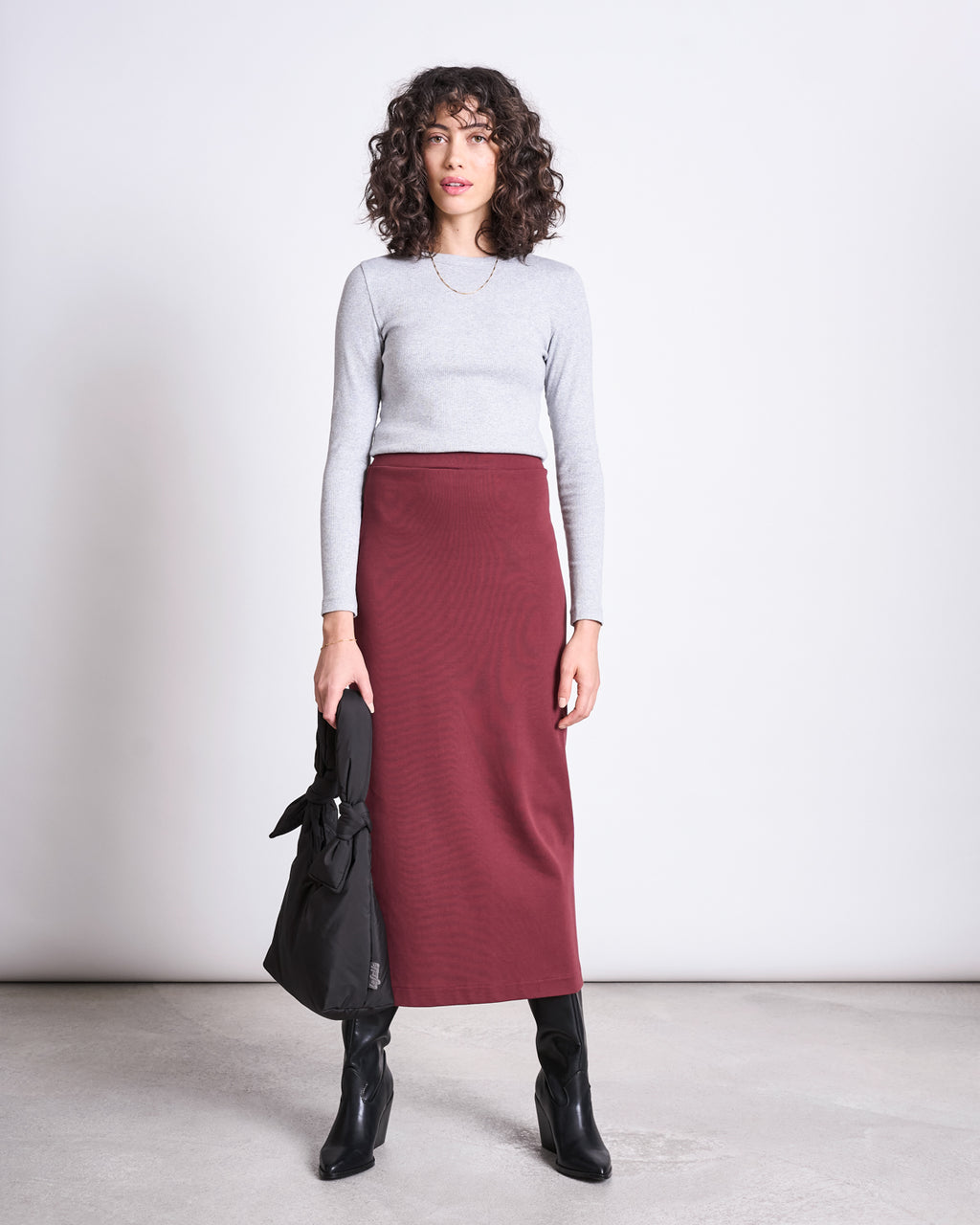 A woman with curly hair wears a fitted light gray long-sleeve top, the jan-n-june MAXI SKIRT BURNITA INTENSE RUST GOTS in maroon, black heeled boots, and holds a black handbag while standing against a plain white background.