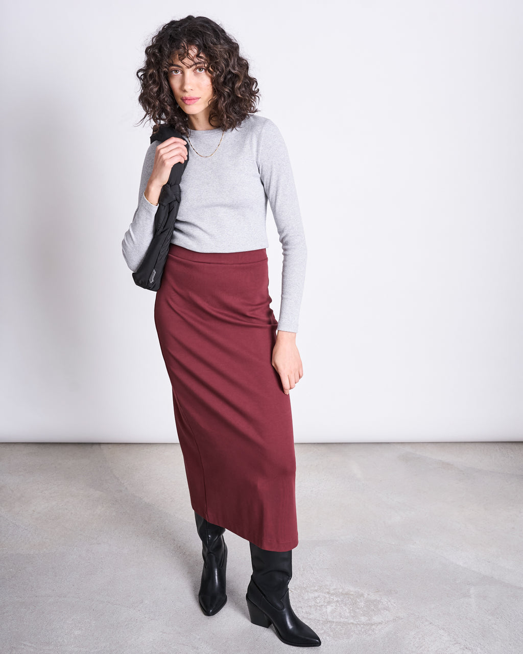 A woman with curly hair wears a light gray long-sleeve top, black boots, and the jan-n-june MAXI SKIRT BURNITA INTENSE RUST GOTS. She holds a black jacket over her shoulder, standing against a plain light gray background.
