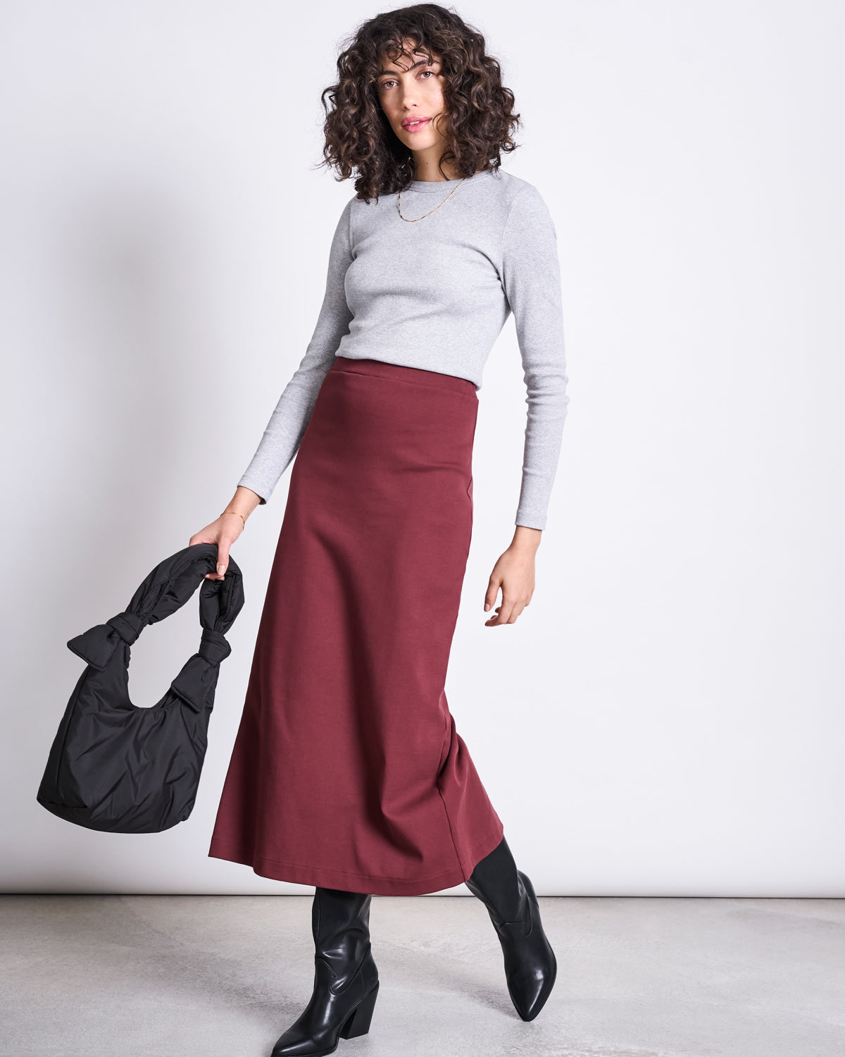 A woman wears the jan-n-june MAXI SKIRT BURNITA INTENSE RUST GOTS with a light gray long-sleeve top and black boots, standing against a plain background and holding a black knotted-handle handbag in her left hand.