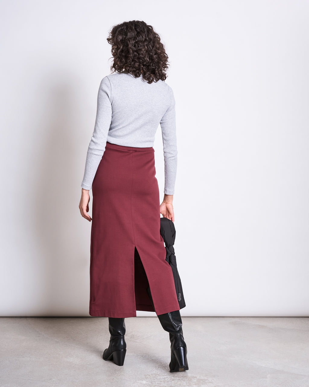 A person with curly hair, wearing a light gray long-sleeve top and the jan-n-june MAXI SKIRT BURNITA INTENSE RUST GOTS, stands facing away holding a black bag and wearing black boots.