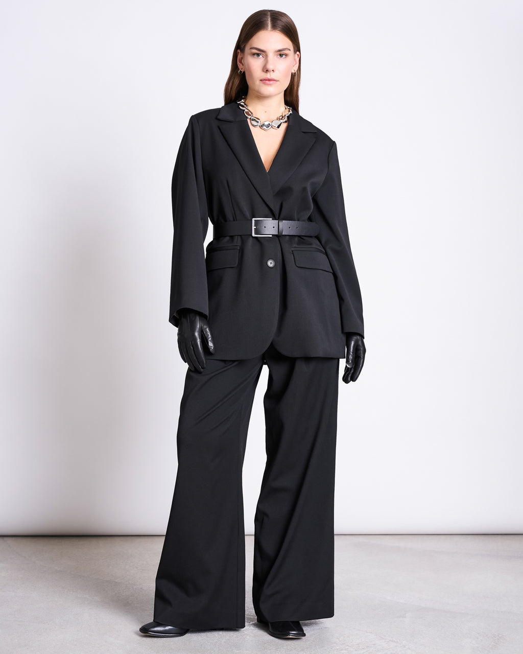 A woman with straight hair and a confident look stands against a plain background wearing the jan-n-june BLAZER DURANCE BLACK, styled with wide-leg trousers, black gloves and shoes, plus a silver necklace.