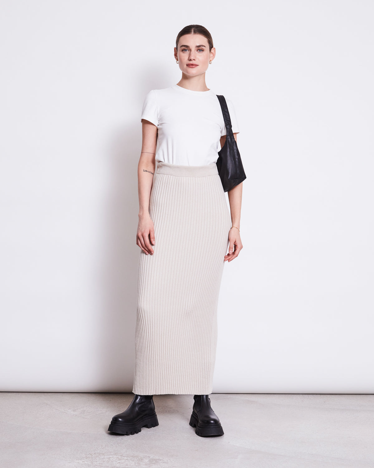 A woman confidently poses against a white wall, wearing the jan-n-june MIDI KNIT SKIRT EBBA IVORY GOTS, a white t-shirt, black chunky boots, and a black shoulder bag. Her hair is tied back as she looks at the camera.