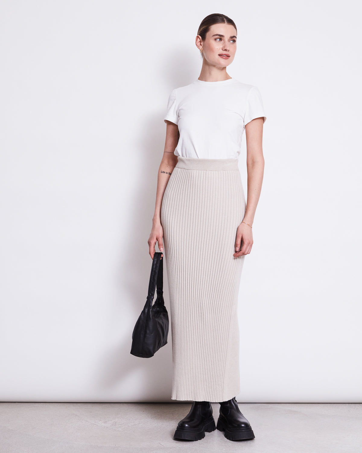 A woman stands against a white background wearing jan-n-junes MIDI KNIT SKIRT EBBA IVORY GOTS, paired with a white organic cotton t-shirt, black chunky shoes, and holding a black bag by her side.