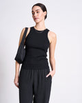 Against a plain white background, a woman wears the jan-n-june ORGANIC COTTON TANK TOP PORTO LONG BLACK GOTS with loose black pants, carries a small black shoulder bag, and has dark hair pulled back.