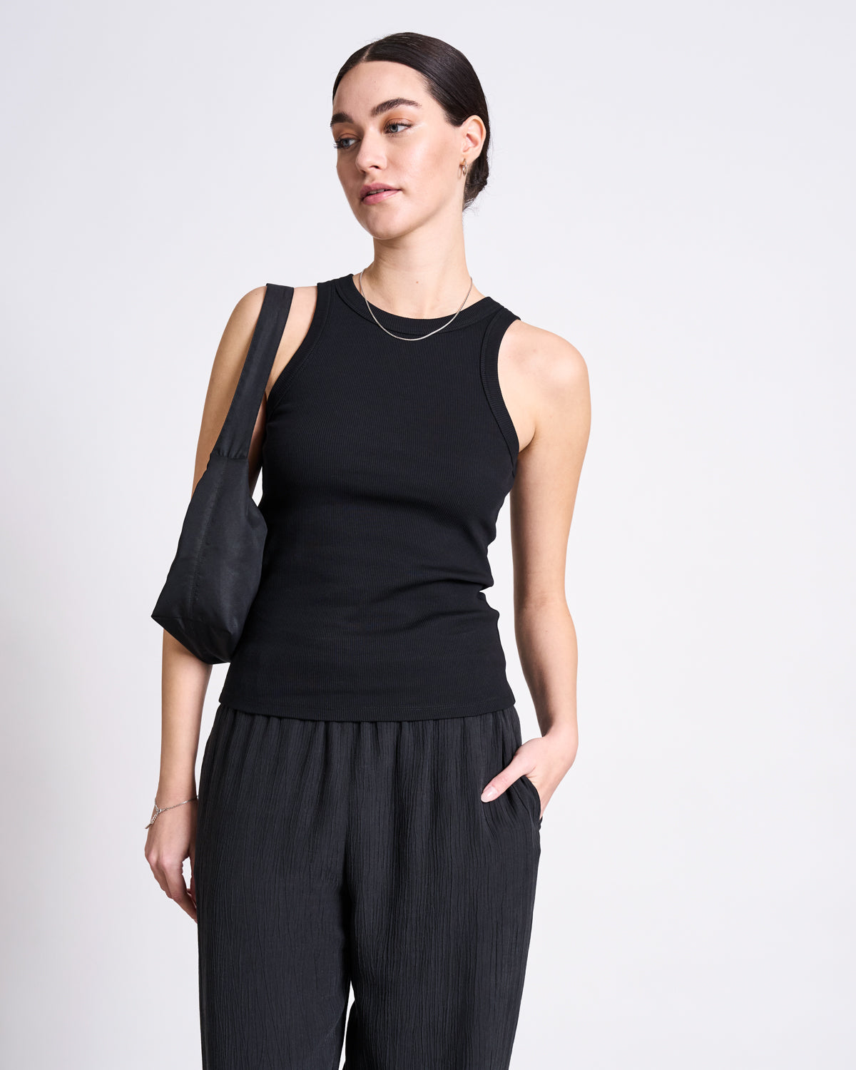Against a plain white background, a woman wears the jan-n-june ORGANIC COTTON TANK TOP PORTO LONG BLACK GOTS with loose black pants, carries a small black shoulder bag, and has dark hair pulled back.