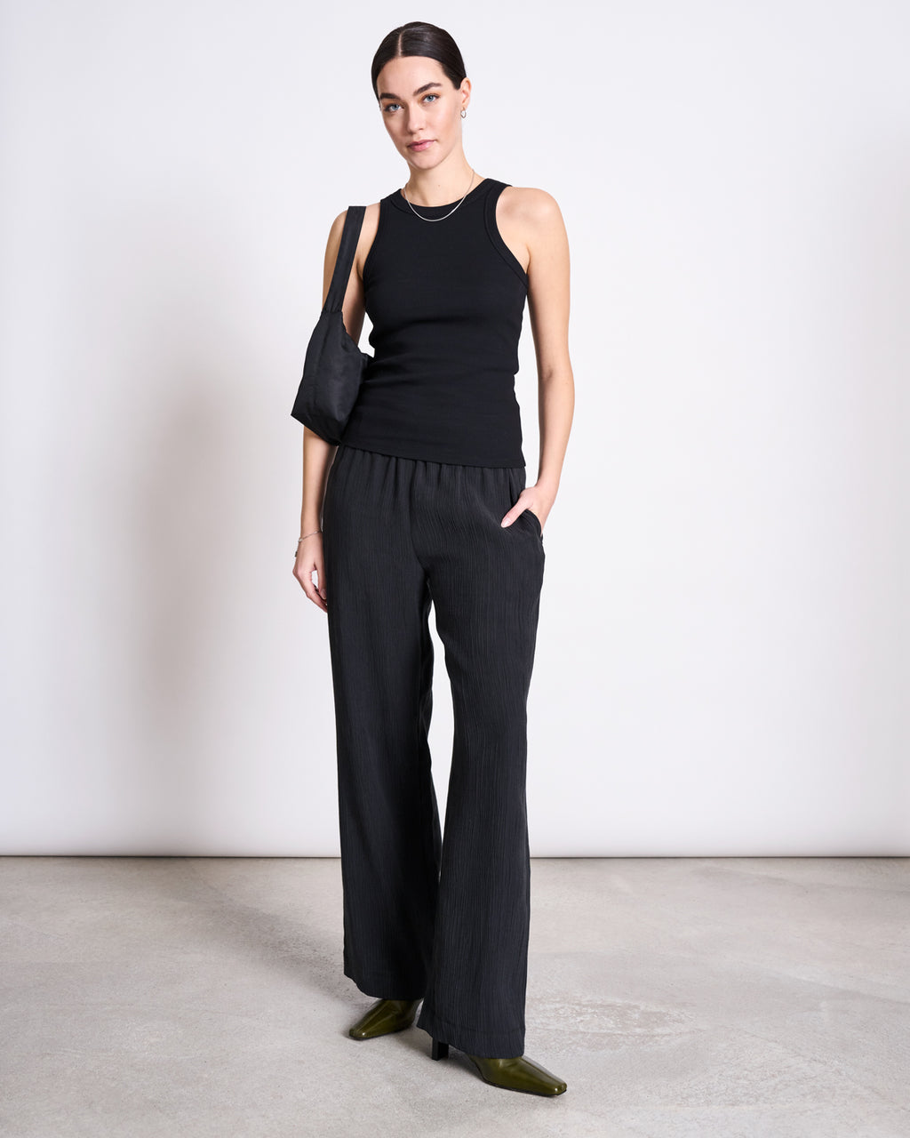 A woman wears the jan-n-june ORGANIC COTTON TANK TOP PORTO LONG BLACK GOTS with loose black pants, green heels, and a black shoulder bag. Her dark hair is pulled back and one hand is in her pocket—a chic look for everyday wear.